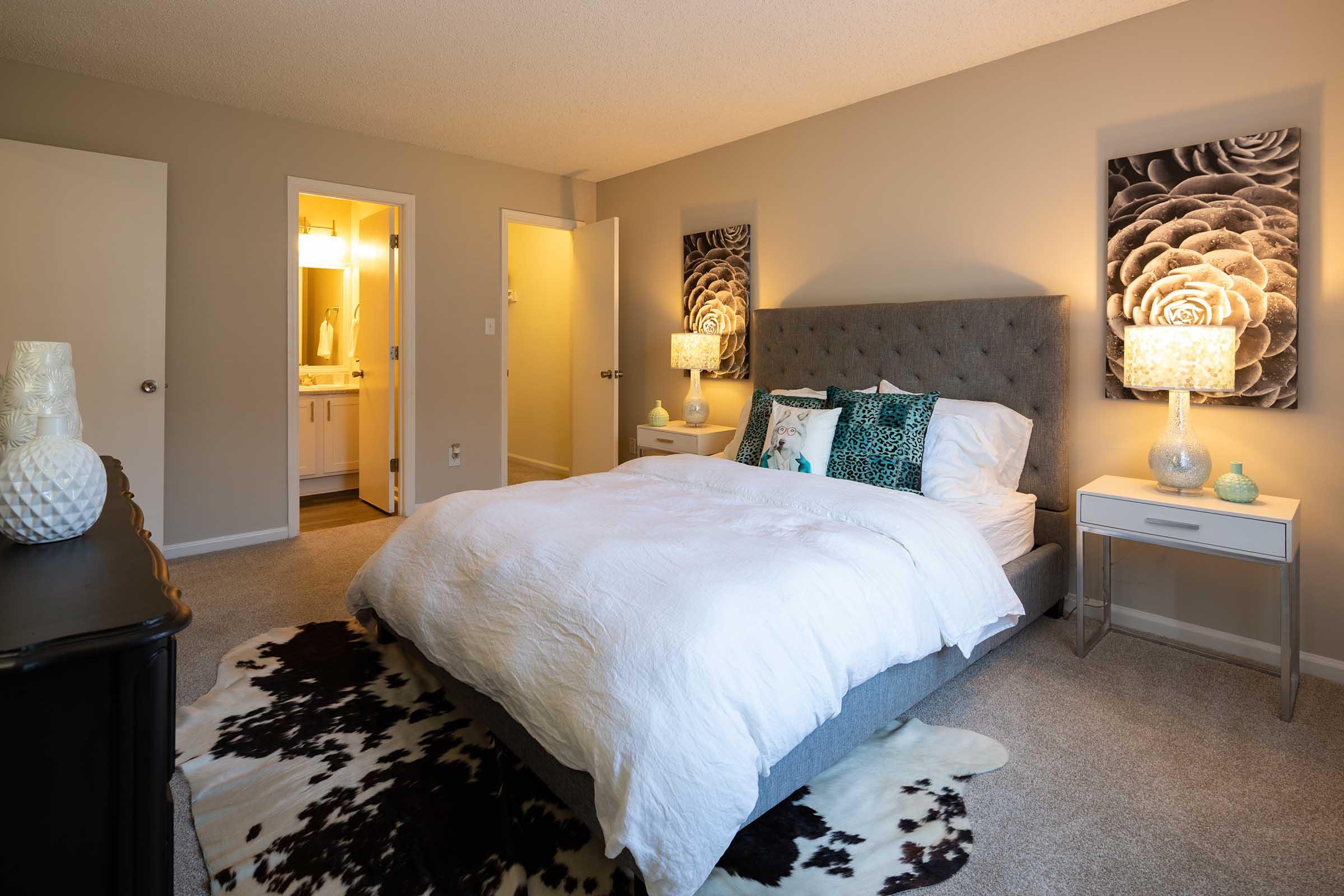 A cozy bedroom featuring a gray upholstered bed with a white comforter and decorative pillows. There are two stylish bedside lamps on either side, and two framed floral wall art pieces. A cowhide rug covers the floor, and a doorway leads to a bathroom, creating a warm and inviting atmosphere.