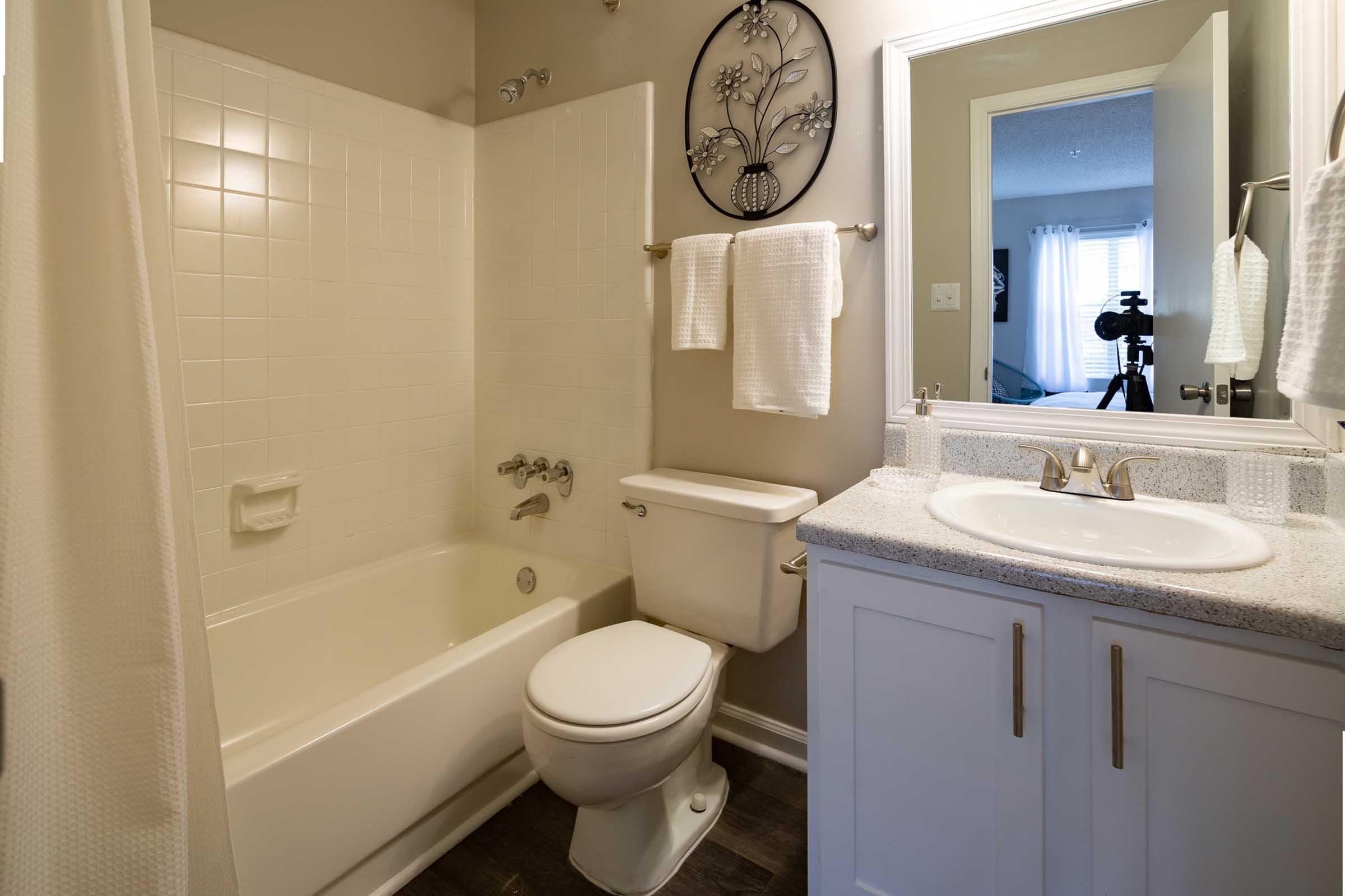 A clean bathroom featuring a white bathtub with a shower curtain, a toilet, and a sink with a granite countertop. White towels hang on towel bars. A mirror reflects part of a room with a window, and a camera is positioned nearby. The walls are painted a neutral color.