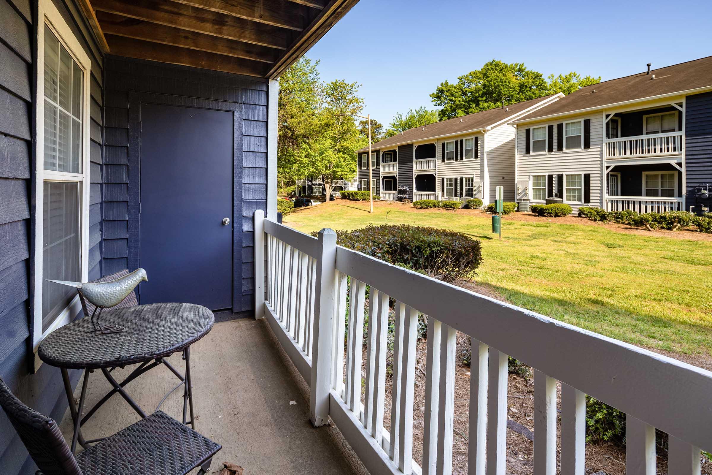 A view from a balcony featuring a small table with a decorative bird and a chair, overlooking a landscaped area with grass and trees. Nearby, a row of apartment buildings can be seen, showcasing a pleasant outdoor living environment.