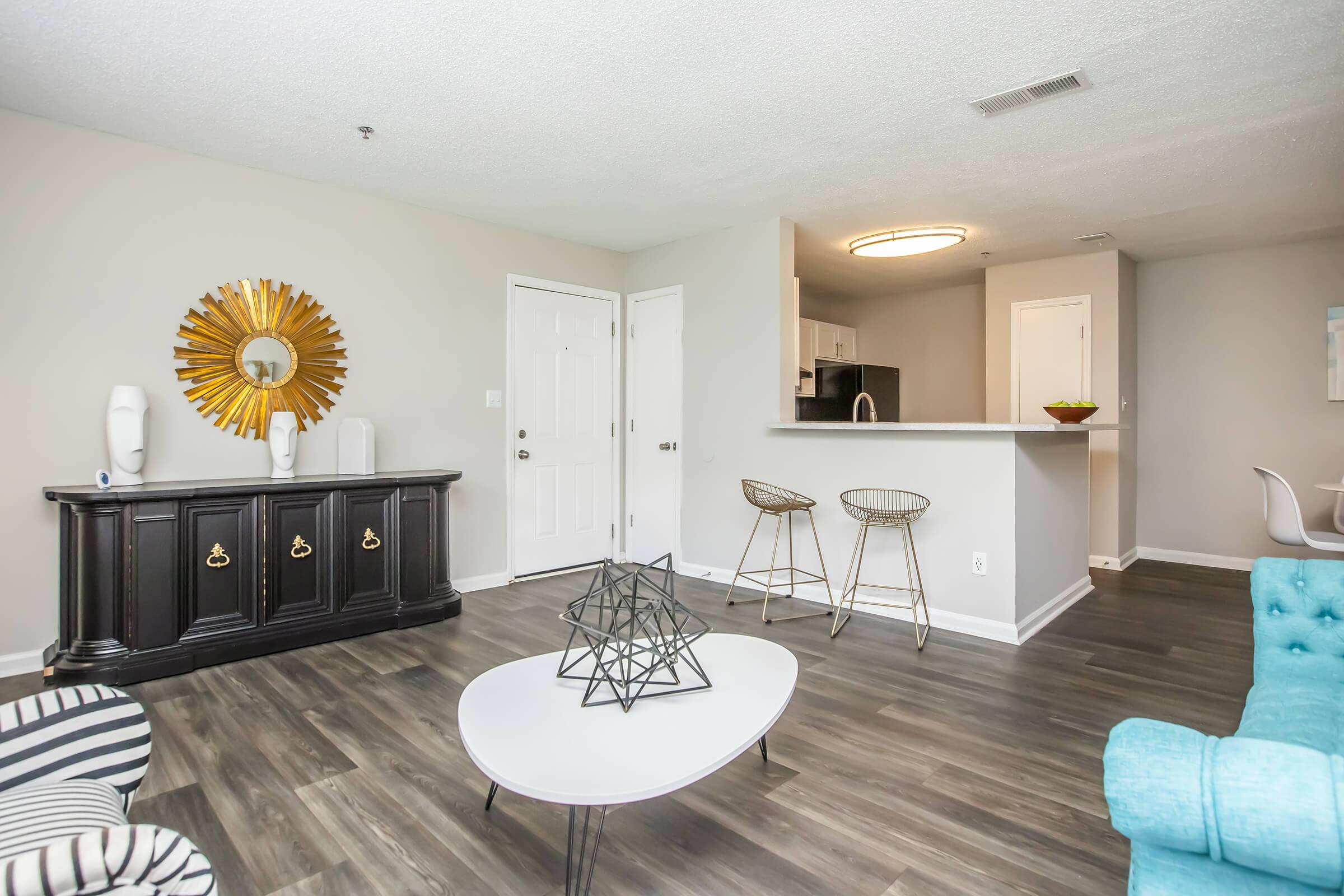 A modern living area featuring a stylish black credenza, a round coffee table with a geometric centerpiece, and two bar stools near a kitchen island. The walls are painted light gray, complemented by a large decorative sunburst mirror. Natural light fills the space, creating a cozy atmosphere.