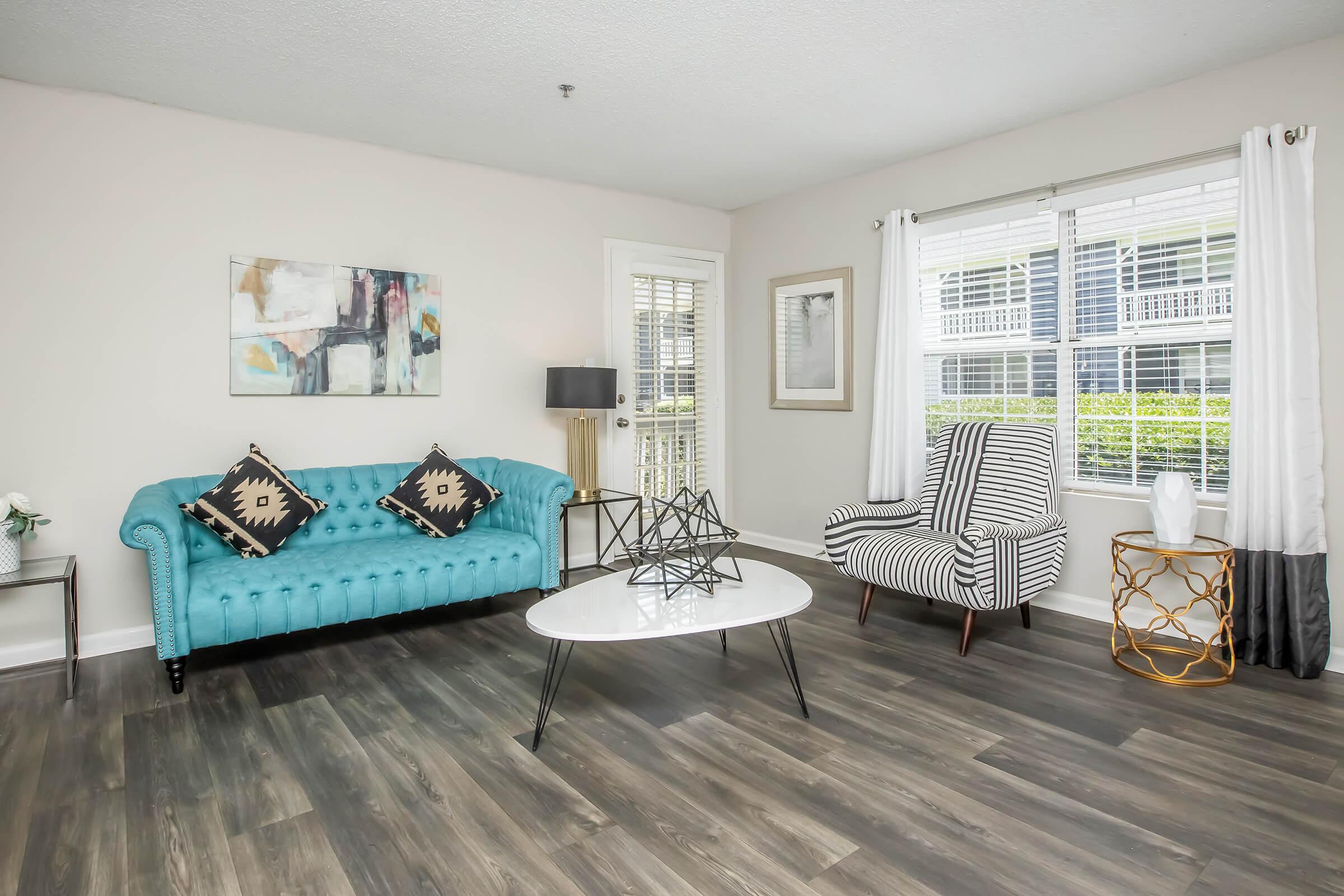 A modern living room featuring a turquoise tufted sofa with black and white decorative pillows, a striped accent chair, a round coffee table with a geometric centerpiece, and large windows allowing natural light. The space has light-colored walls and a sleek laminate floor, creating a stylish and inviting atmosphere.