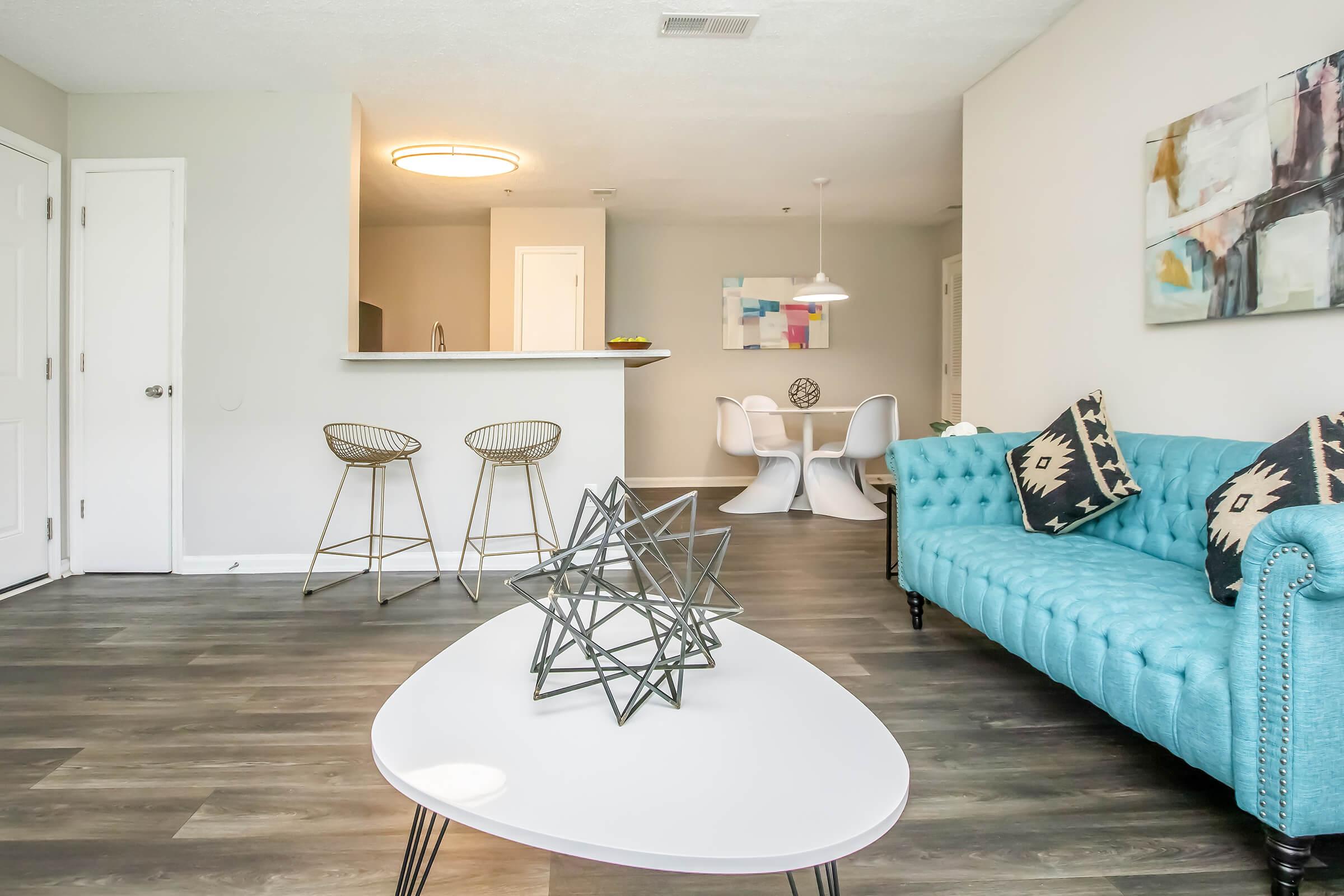 A modern living room featuring a teal tufted sofa with patterned throw pillows, a white circular coffee table with a geometric centerpiece, and bar stools at a kitchen counter. In the background, there is a dining area with a round table and white chairs, complemented by contemporary artwork on the wall.