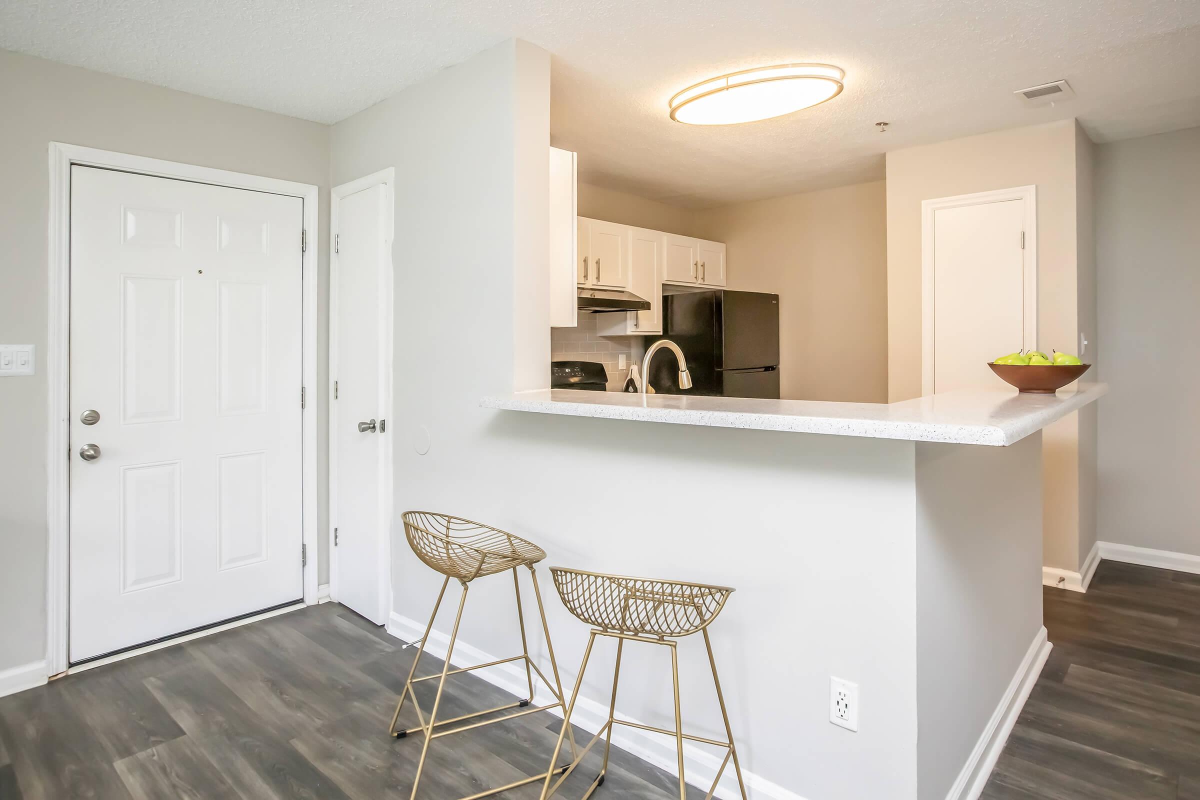 A modern kitchen area featuring a small breakfast bar with two gold metal stools, a white countertop, and a view into a well-lit kitchen equipped with white cabinets and black appliances. The walls are painted in a light gray, creating a contemporary and inviting atmosphere.