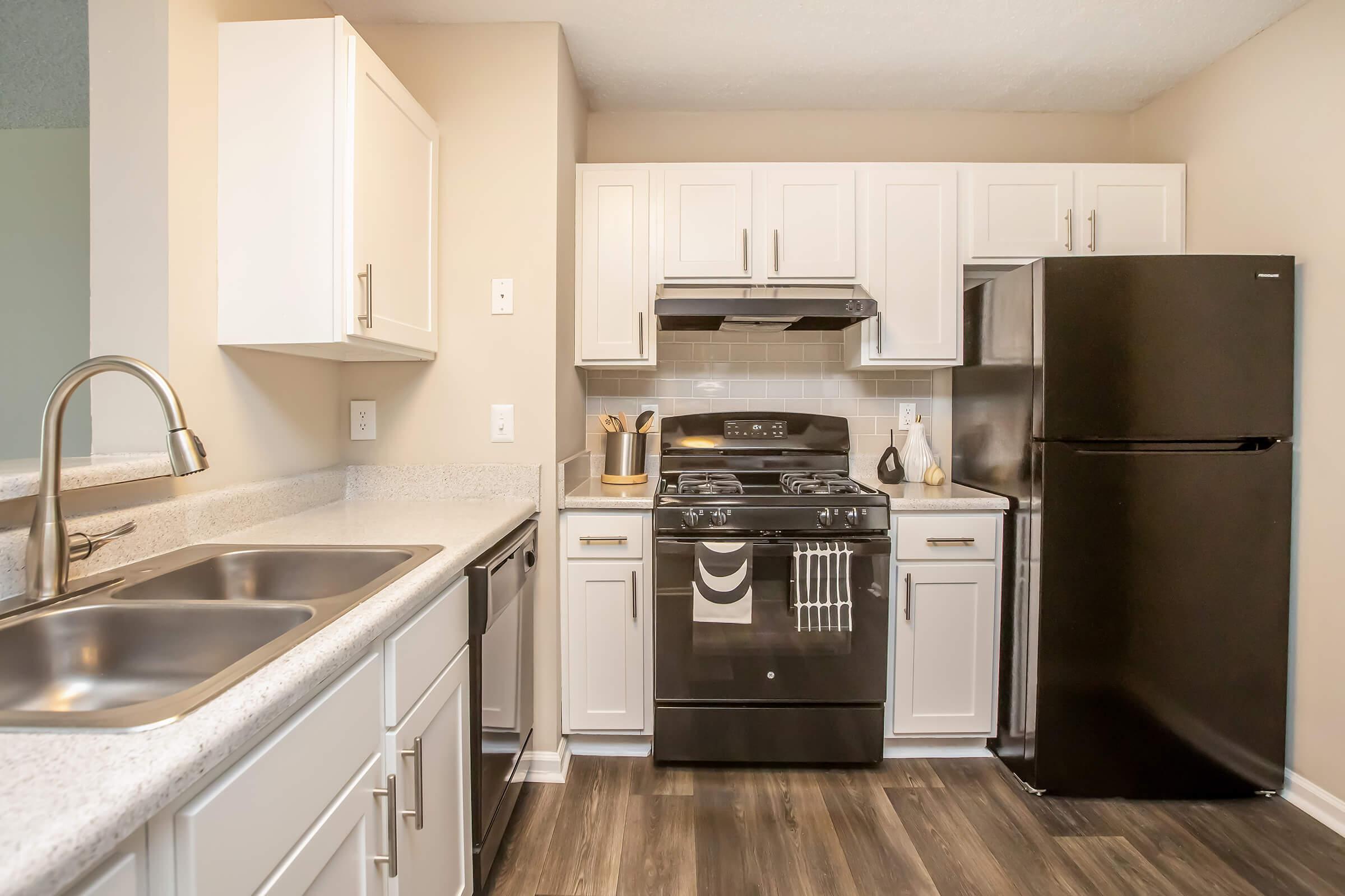 A modern kitchen featuring white cabinets, a black refrigerator, a gas stove with an overhead exhaust fan, and dual sinks. The countertops are light-colored, and the flooring is a dark wood laminate, creating a contemporary and stylish cooking space.