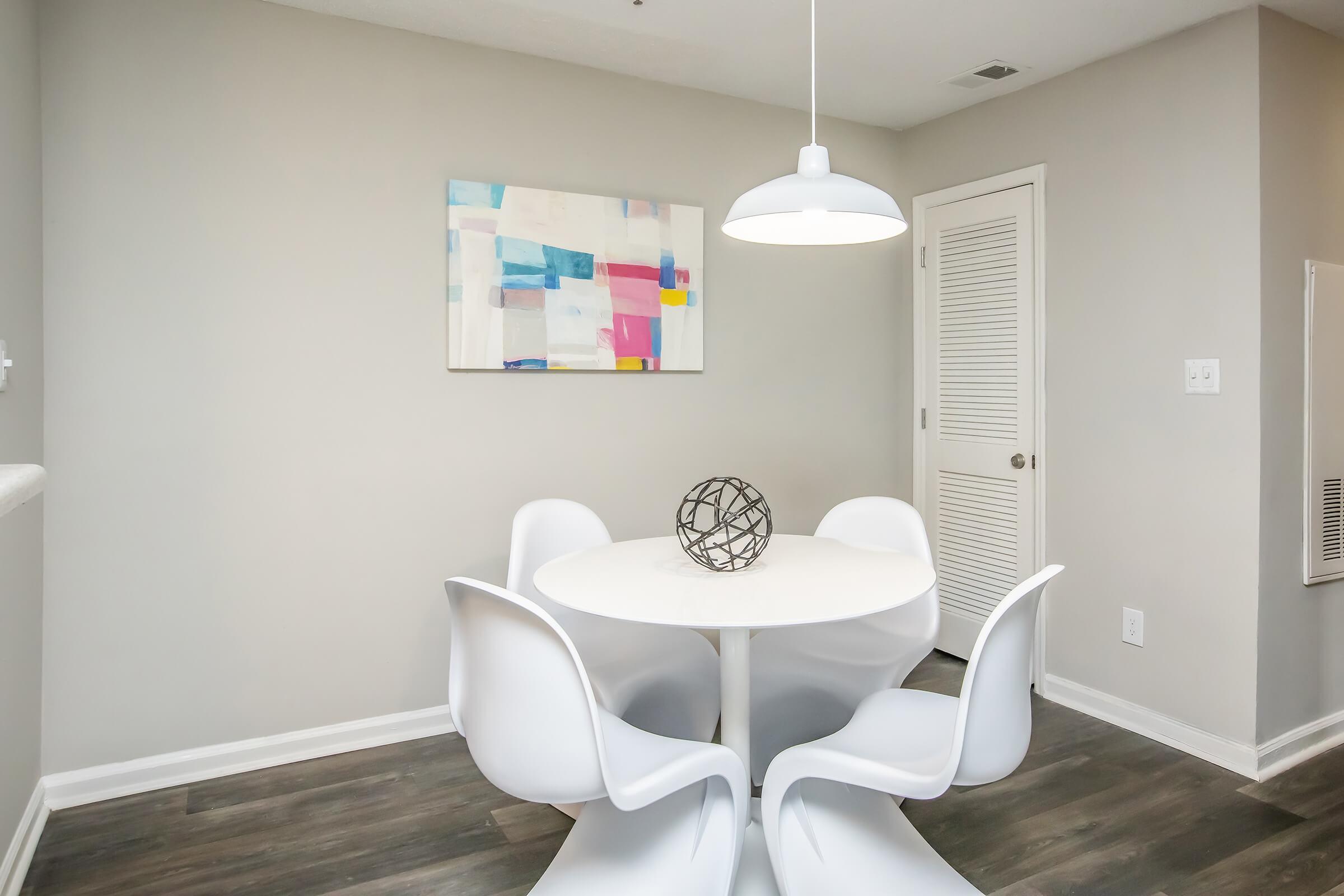A modern dining area featuring a round white table surrounded by four white chairs. Above the table hangs a minimalistic pendant light. The wall is adorned with a colorful abstract painting, and a decorative sphere sits on the table. The floor has a wood-like finish, and the walls are painted in a light neutral color.
