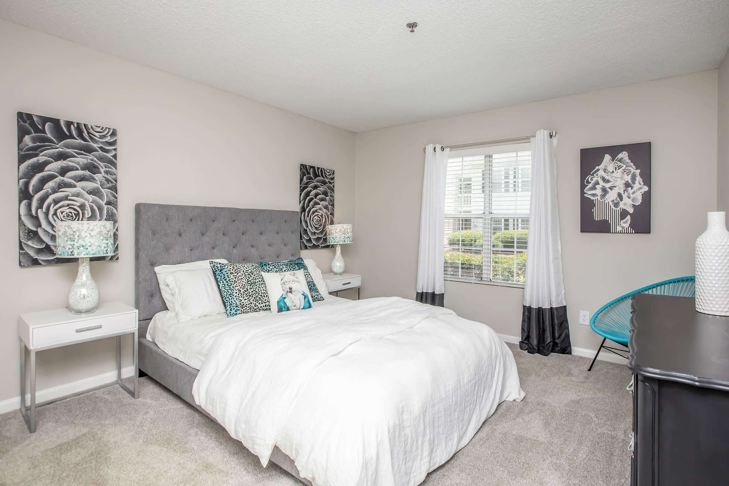 A modern bedroom featuring a gray upholstered bed with white bedding and decorative pillows. The walls are adorned with floral artwork, and there are two bedside tables with lamps. A large window lets in natural light, showcasing a neatly kept outdoor view. A stylish accent chair and a dark dresser complete the serene décor.