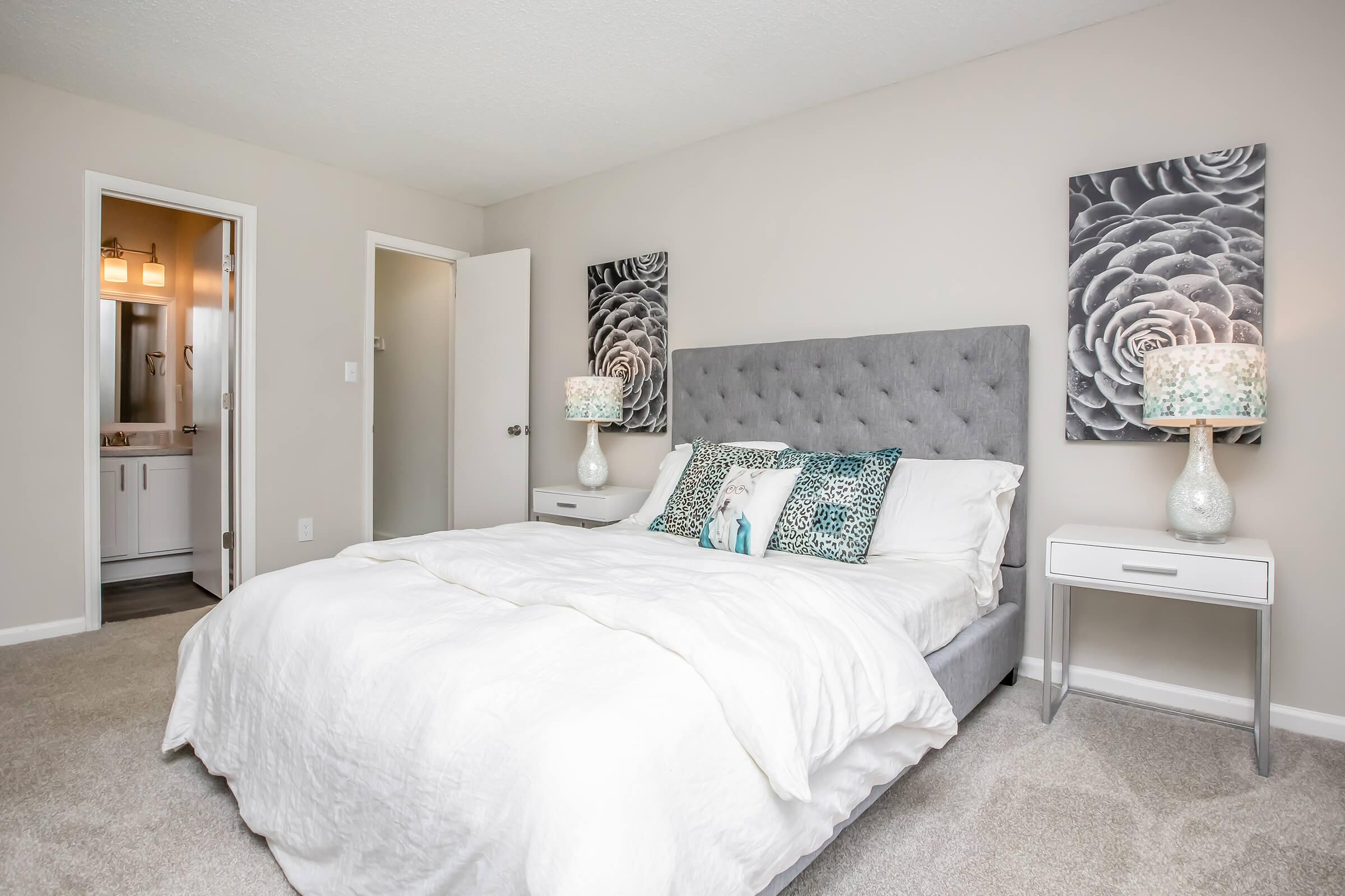 A modern bedroom featuring a gray upholstered bed with white bedding and decorative pillows. On either side of the bed are matching nightstands with lamps. The walls are a soft beige color, adorned with two patterned wall art pieces. A doorway leads to a bathroom in the background.