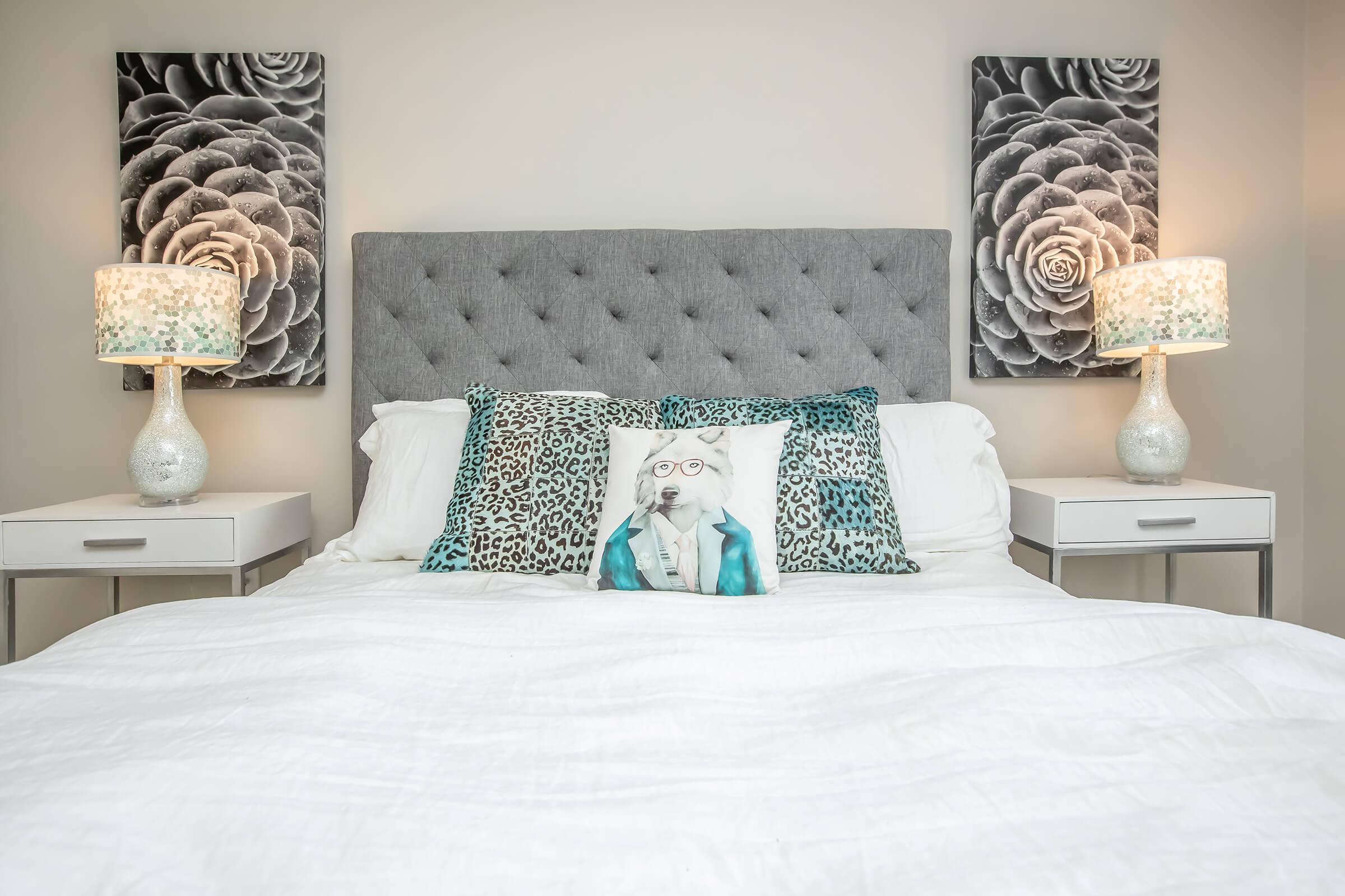 A modern bedroom featuring a gray tufted headboard, white bedding, and decorative pillows. On either side of the bed are white nightstands with stylish lamps. The wall art includes black and white floral images, creating a chic and inviting atmosphere.