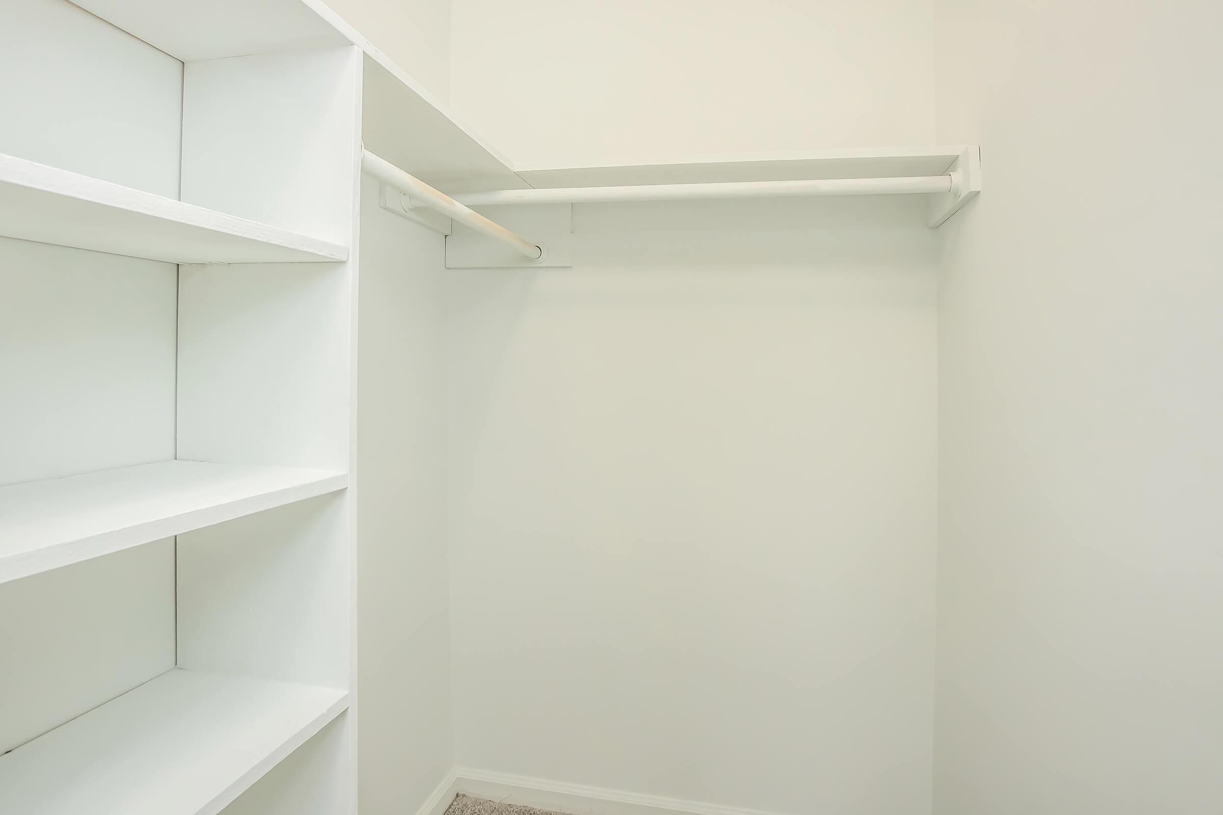 Empty closet space with white shelves on the left and a clothing rod along the back wall. The walls and shelves are painted white, and the floor is carpeted in a light color. The overall appearance is clean and minimalistic.