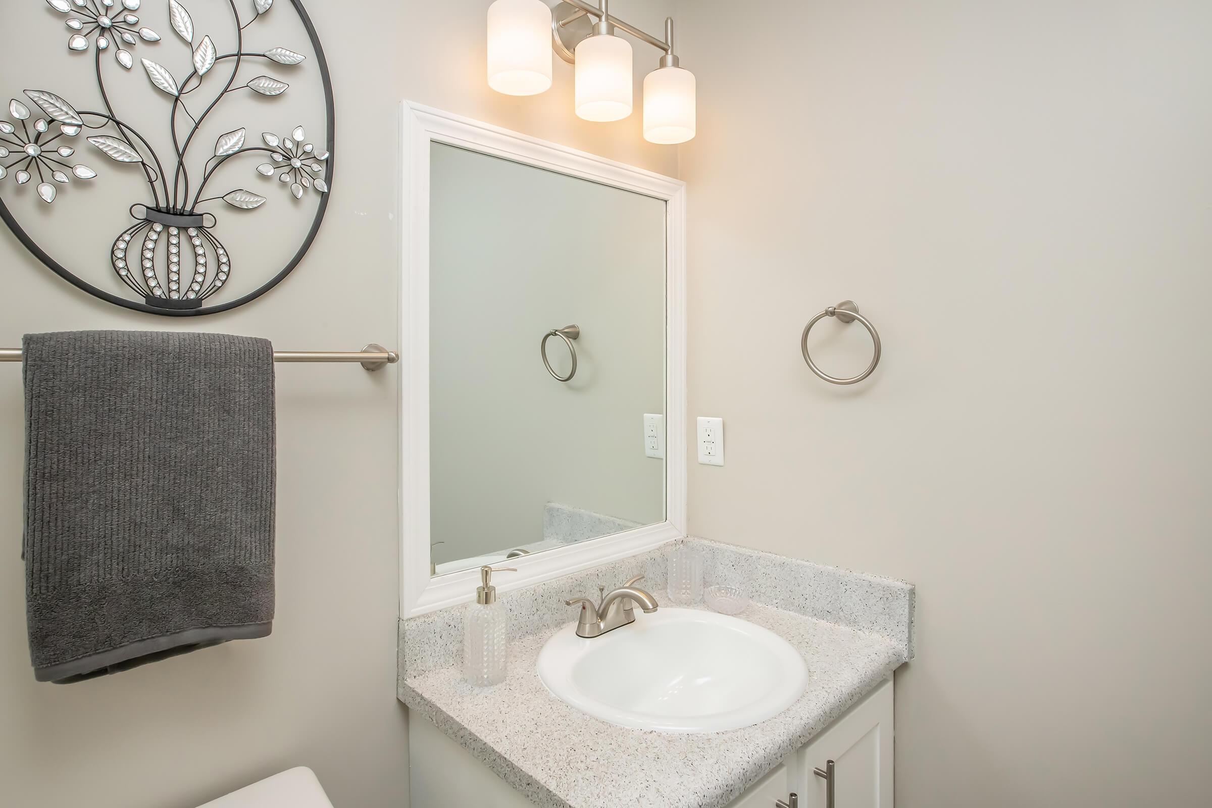 A modern bathroom with a light-colored wall, featuring a white sink with a faucet and a circular mirror above it. A towel hangs on a rack nearby, and a wall-mounted towel ring is visible. The decor includes an artistic wall piece of a vase with flowers, complemented by warm lighting from three hanging fixtures.