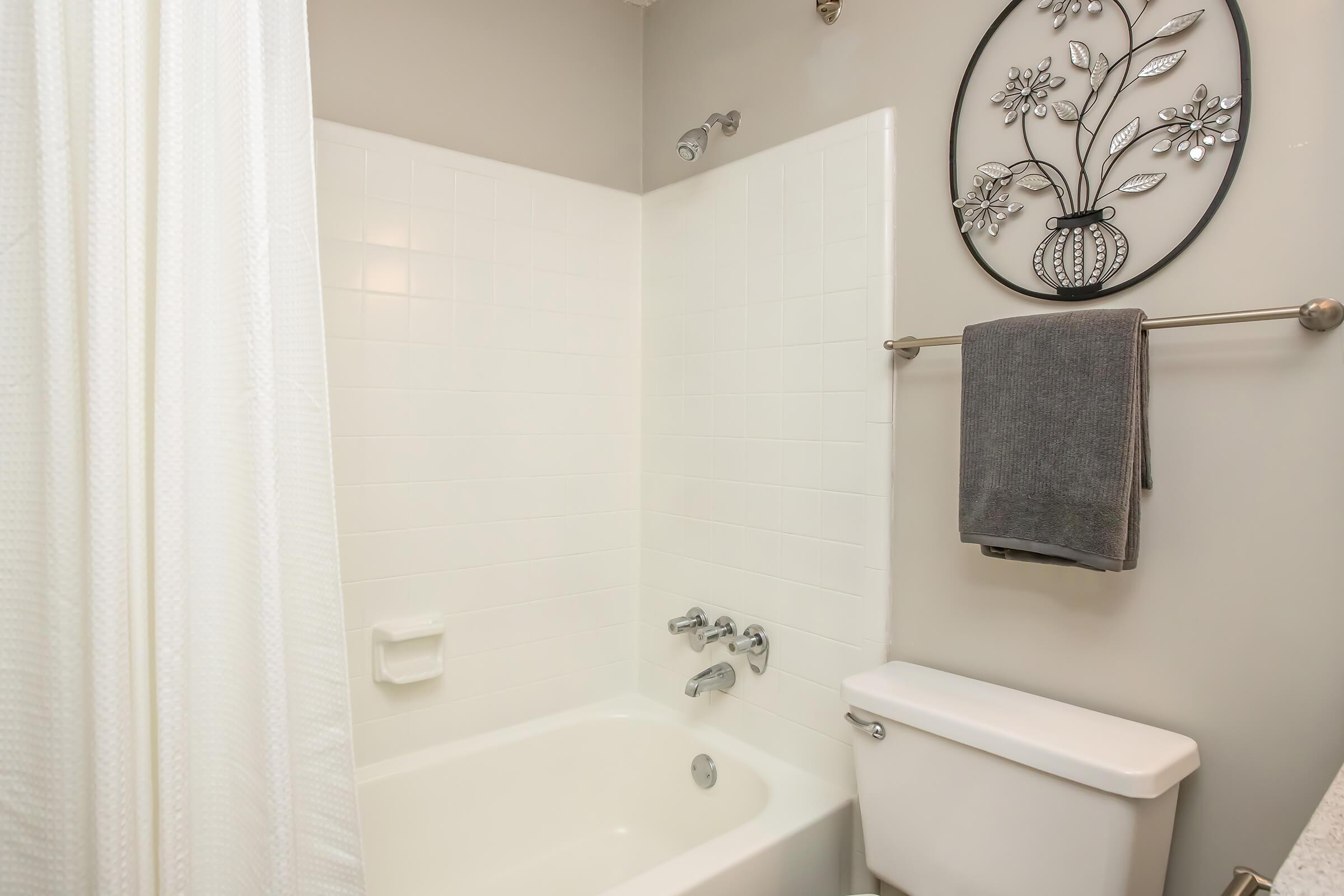 A clean, modern bathroom featuring a white bathtub with shower fixtures, a decorative wall hanging of flowers in a vase, a towel rack with a gray towel, and a white toilet. The walls are light-colored, and a shower curtain partially covers the bathtub.