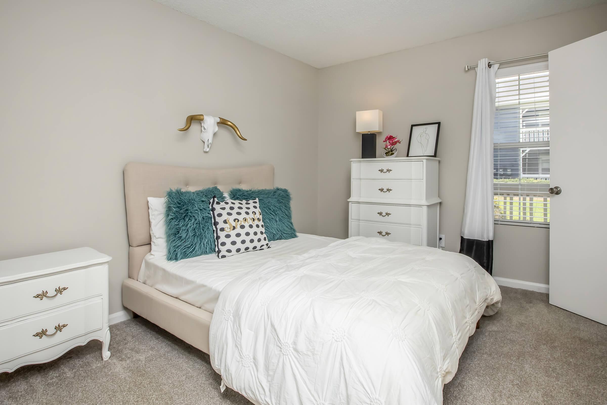 Cozy bedroom featuring a beige upholstered bed with decorative pillows, a white comforter, and a small nightstand. A white dresser with gold accents stands nearby, and a bull skull mounted on the wall adds a rustic touch. Natural light filters through a window with sheer curtains.