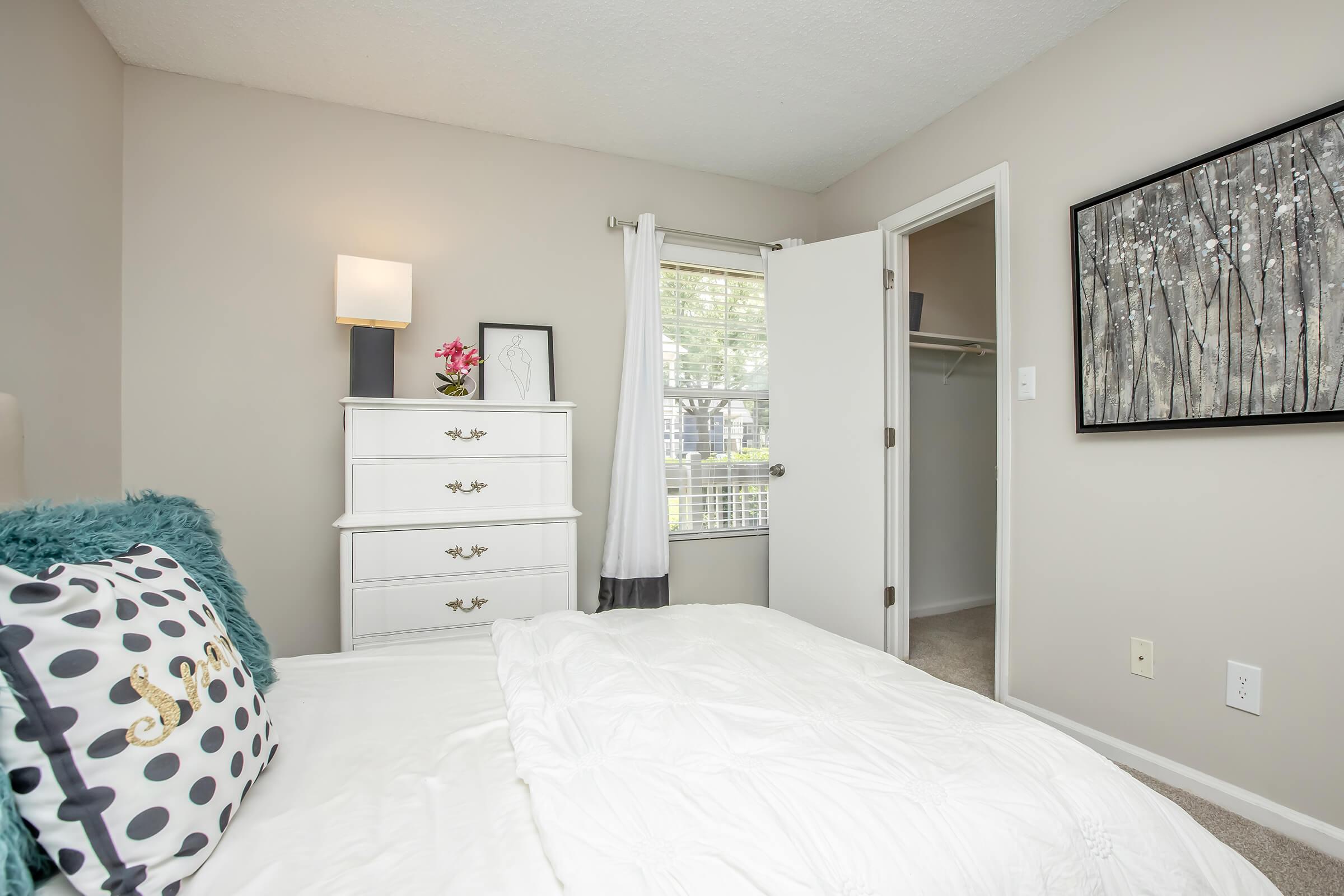 A cozy bedroom featuring a neatly made bed with a white comforter and decorative pillow, a white dresser against the wall, a lamp, a framed art piece, and a closet door. Natural light filters in through a window adorned with sheer curtains.