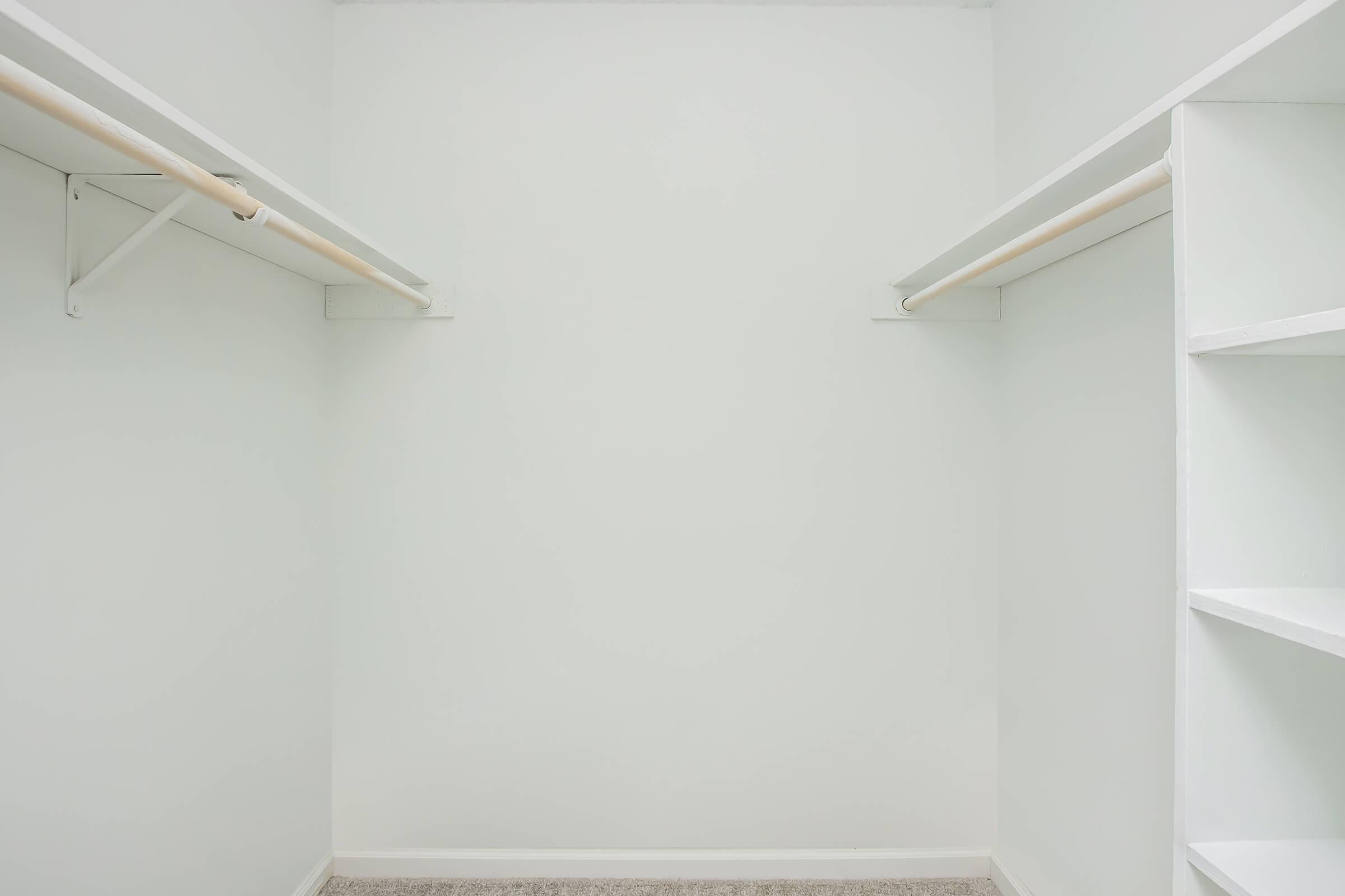 A clean, empty walk-in closet with white walls and light beige carpet. Two hanging rods are mounted on opposite sides, and a single shelf is visible on the right side. The overall appearance is bright and minimalist, ideal for storage or organization.