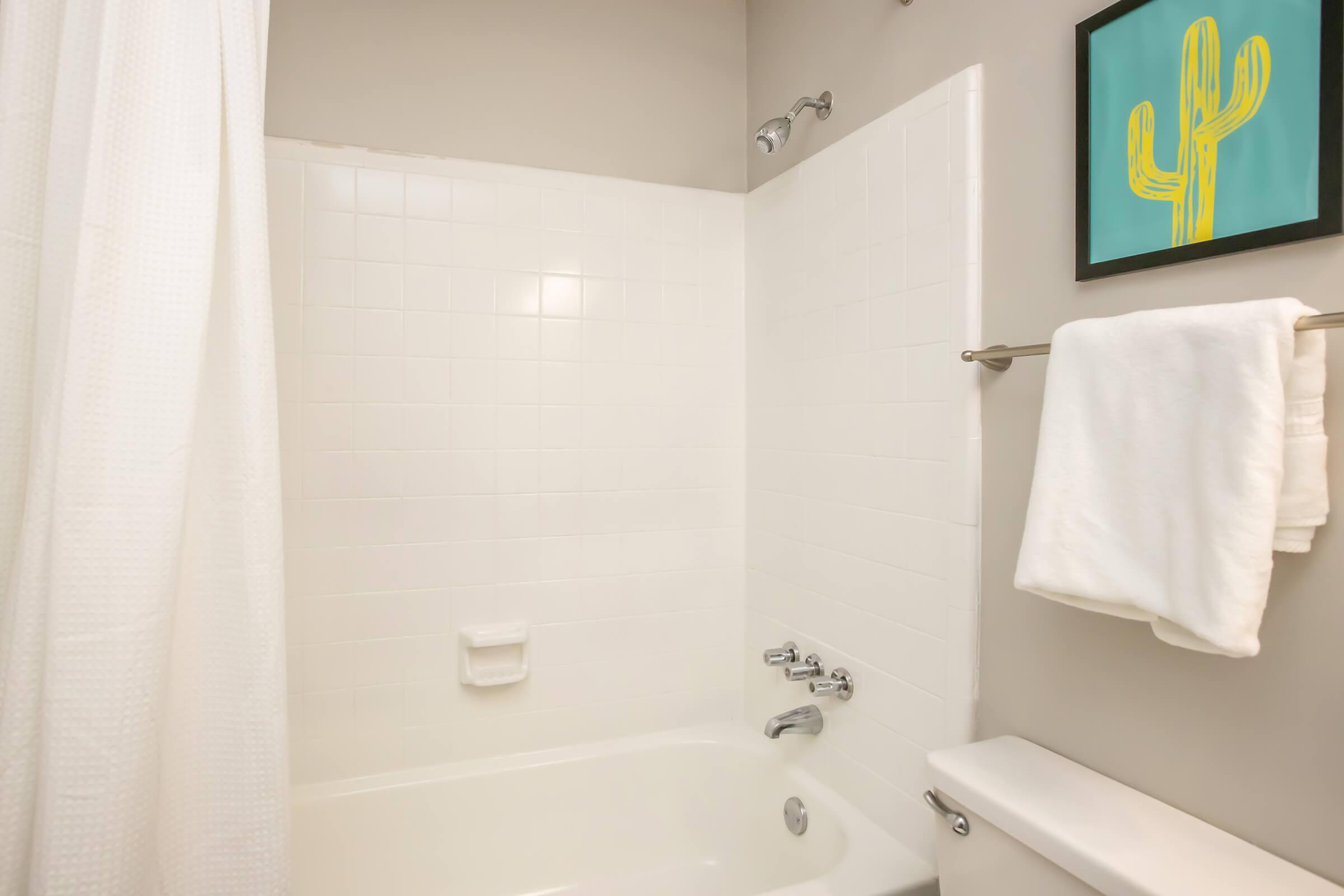 A clean and minimalist bathroom featuring a white bathtub and shower with a curtain. The walls are tiled in white, and there's a pale-colored towel hanging on a bar. A framed artwork of a cactus hangs on the wall, adding a pop of color to the space.