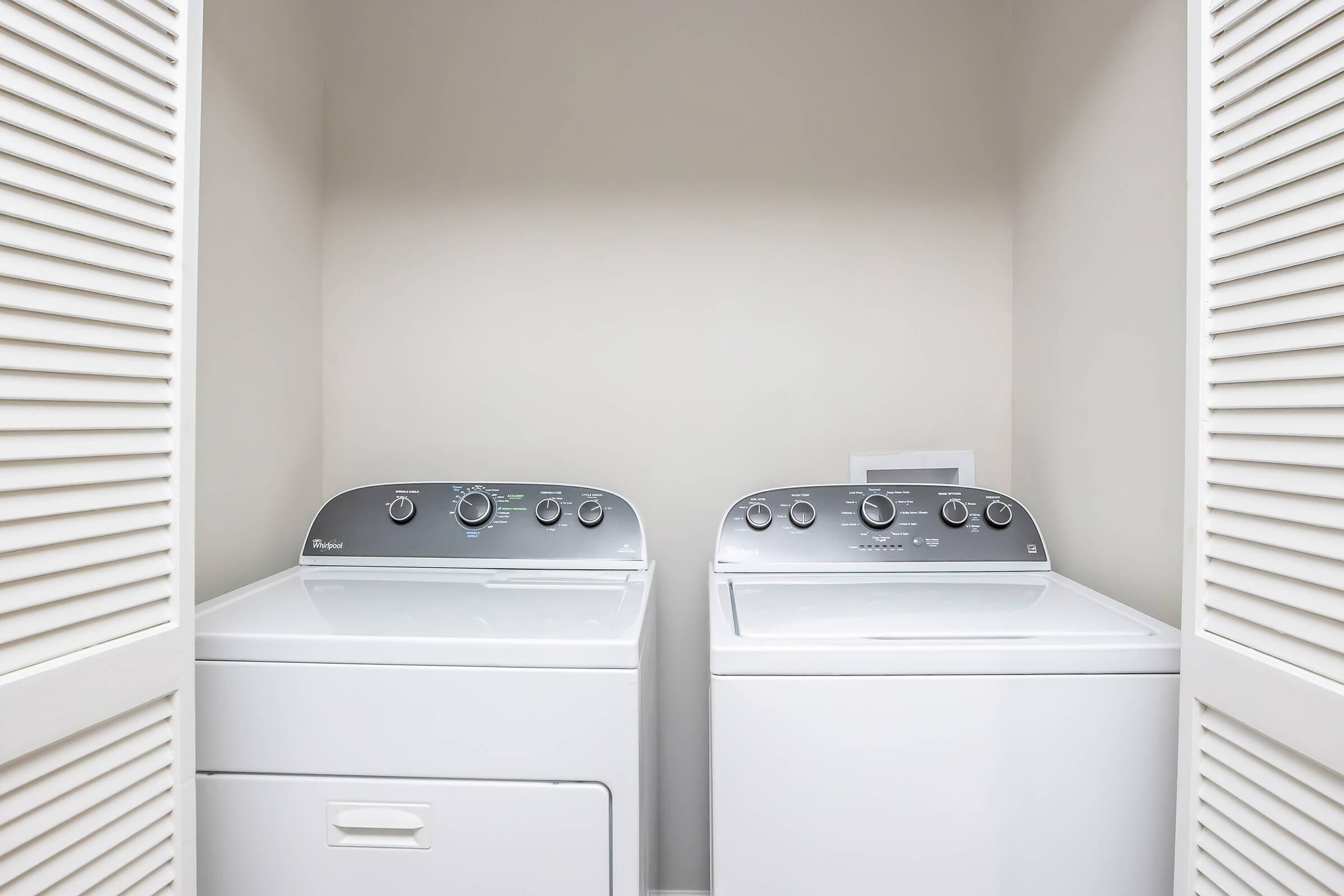A washing machine and dryer are placed side by side in a laundry closet. Both appliances feature modern controls and a clean, white design. The space is well-organized with closed louvered doors, and the walls are painted a neutral color, creating a functional and tidy laundry area.