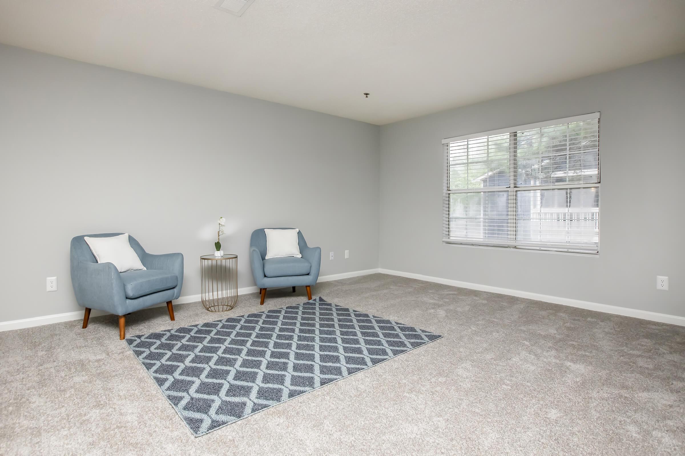 A cozy living room featuring two light blue armchairs with white cushions, a small round table between them, and a patterned area rug on beige carpet. A window with blinds allows natural light to enter the space, which is painted in a soft gray tone.