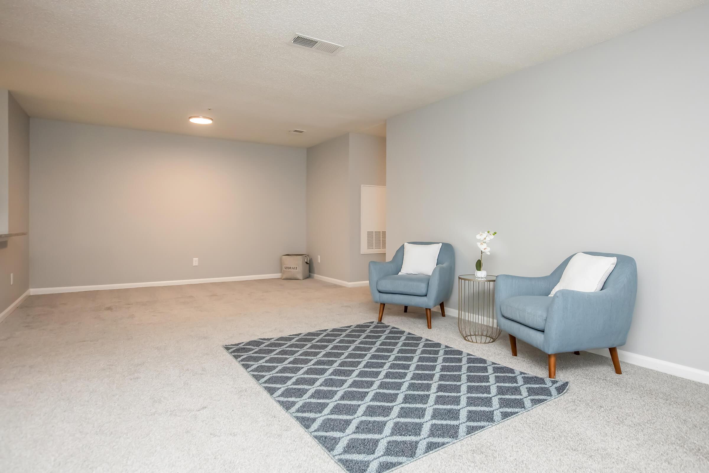 A well-lit, empty living room featuring light gray walls and plush carpet. Two blue armchairs with white cushions are positioned on either side of a decorative area rug with a geometric pattern. A small round table with a vase of flowers is nearby, creating a cozy atmosphere.