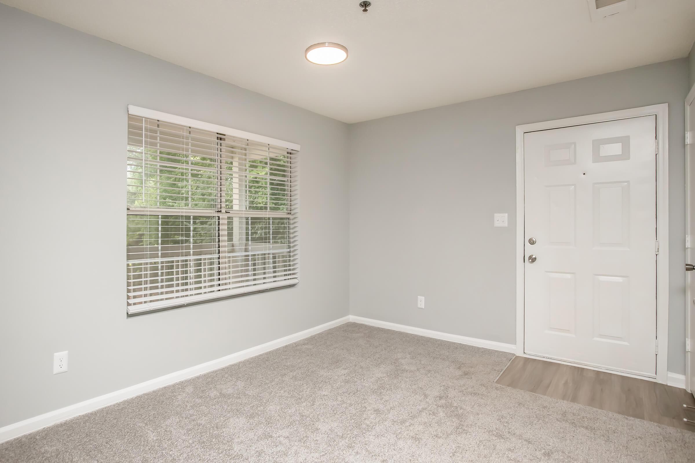 A light gray room with new carpet and a window featuring white blinds. There's a white door on the right, and the wall has a light fixture. The space appears empty and well-lit, creating a clean and inviting atmosphere.