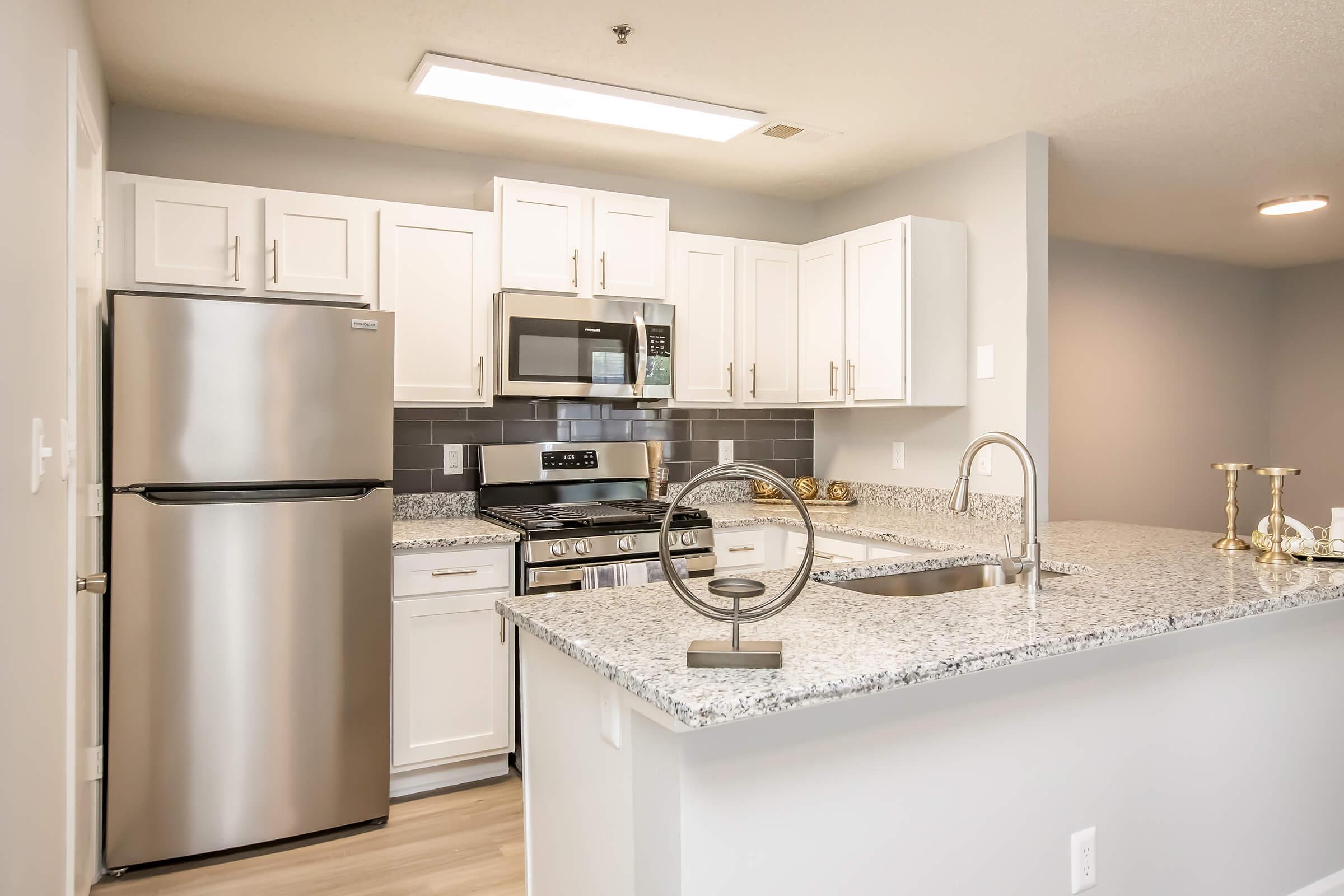Modern kitchen featuring stainless steel appliances, including a refrigerator, microwave, and oven, with white cabinetry and granite countertops. The kitchen is well-lit with a ceiling light and has an open layout. A decorative piece is placed on the countertop, enhancing the contemporary design.