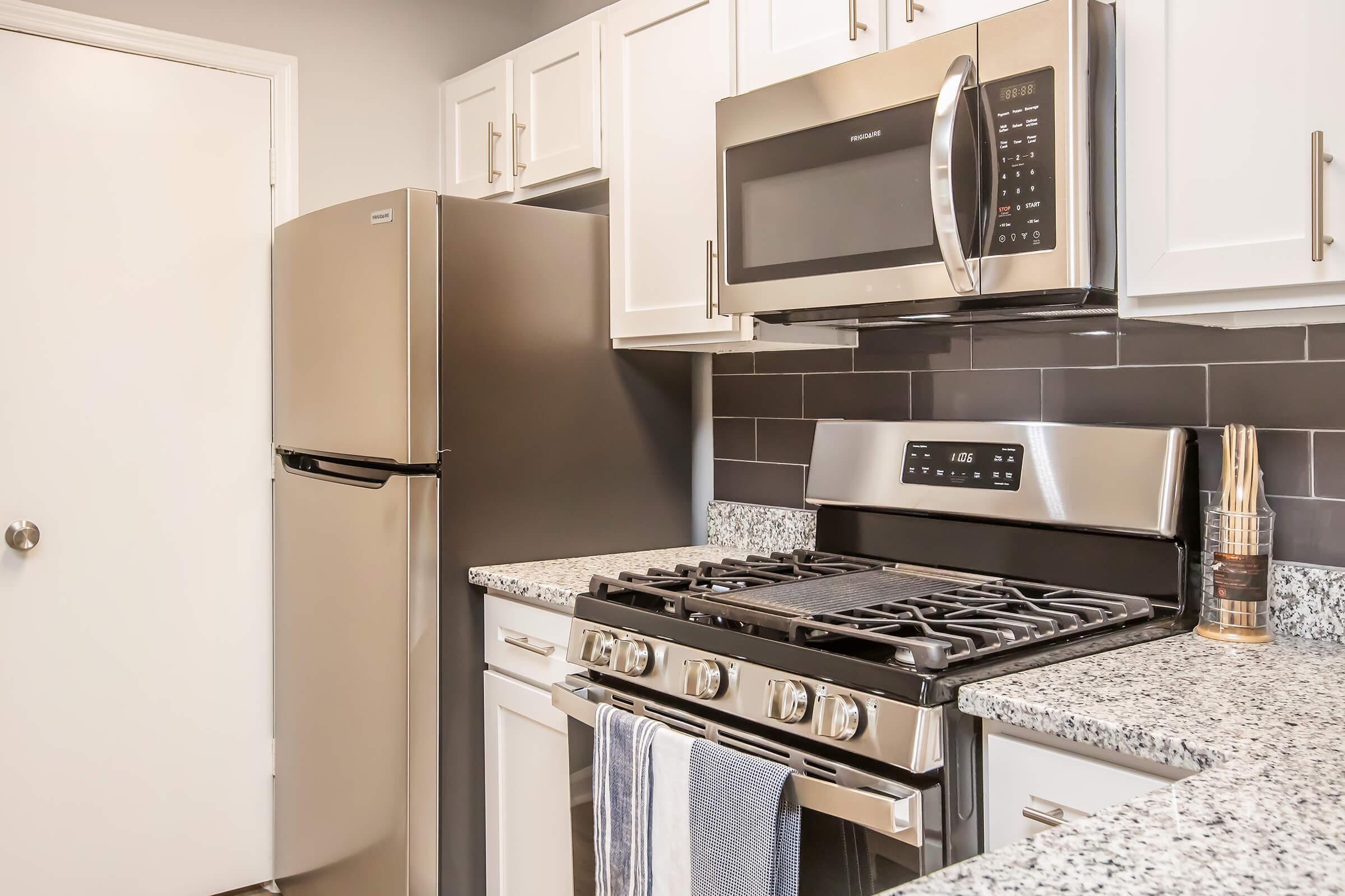 Modern kitchen featuring stainless steel appliances, including a microwave and gas stove, with a granite countertop. The refrigerator is to the left, and there are light-colored cabinets above and below. The back splash is dark, adding contrast to the overall bright and contemporary design.