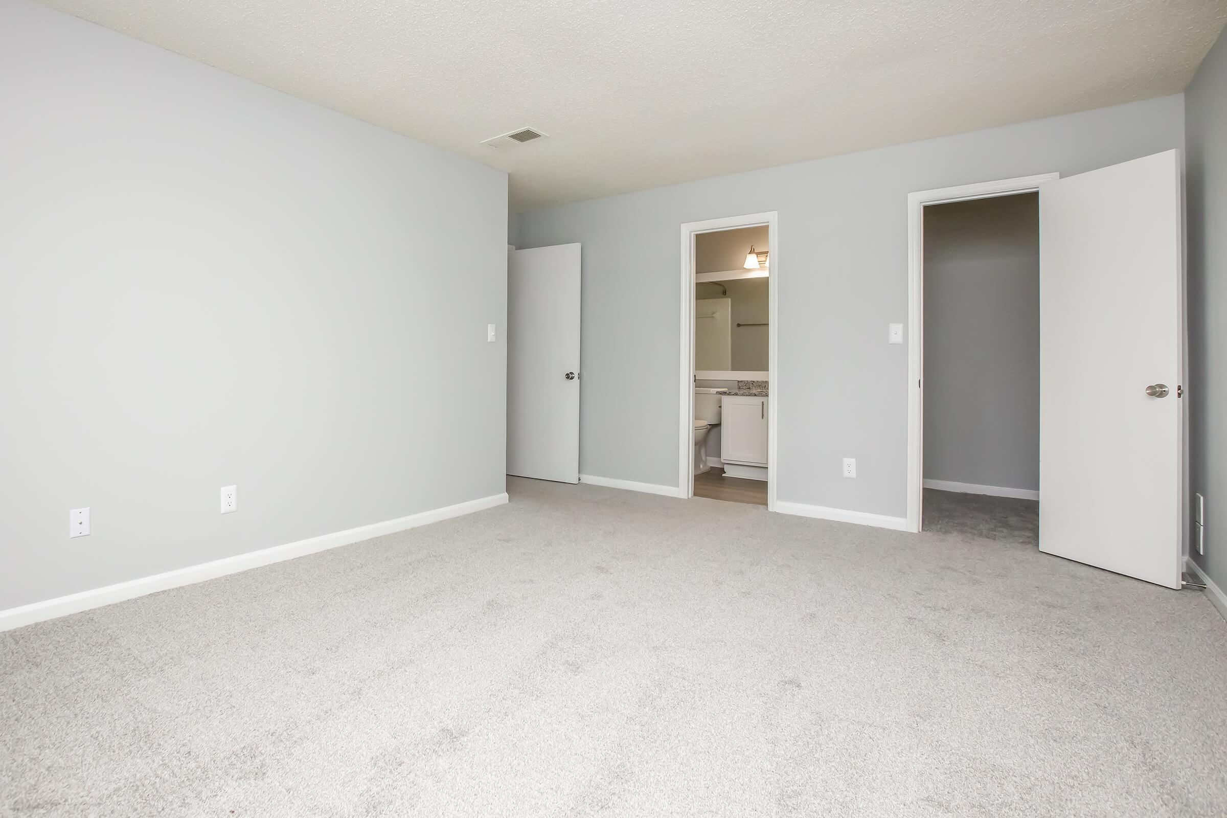 Empty room with light gray walls and beige carpet. Two doors lead to adjacent spaces, one partially visible bathroom area. The room is well-lit with natural light, offering a spacious and clean appearance, suitable for various uses.