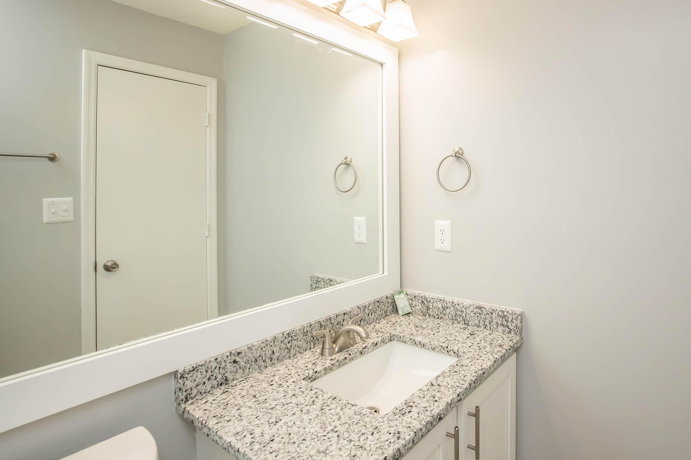 A modern bathroom featuring a granite countertop with a sink, a large mirror above, and a round towel ring mounted on the wall. The walls are painted light gray, and there is a door visible, leading to another space. The overall design is clean and minimalistic.
