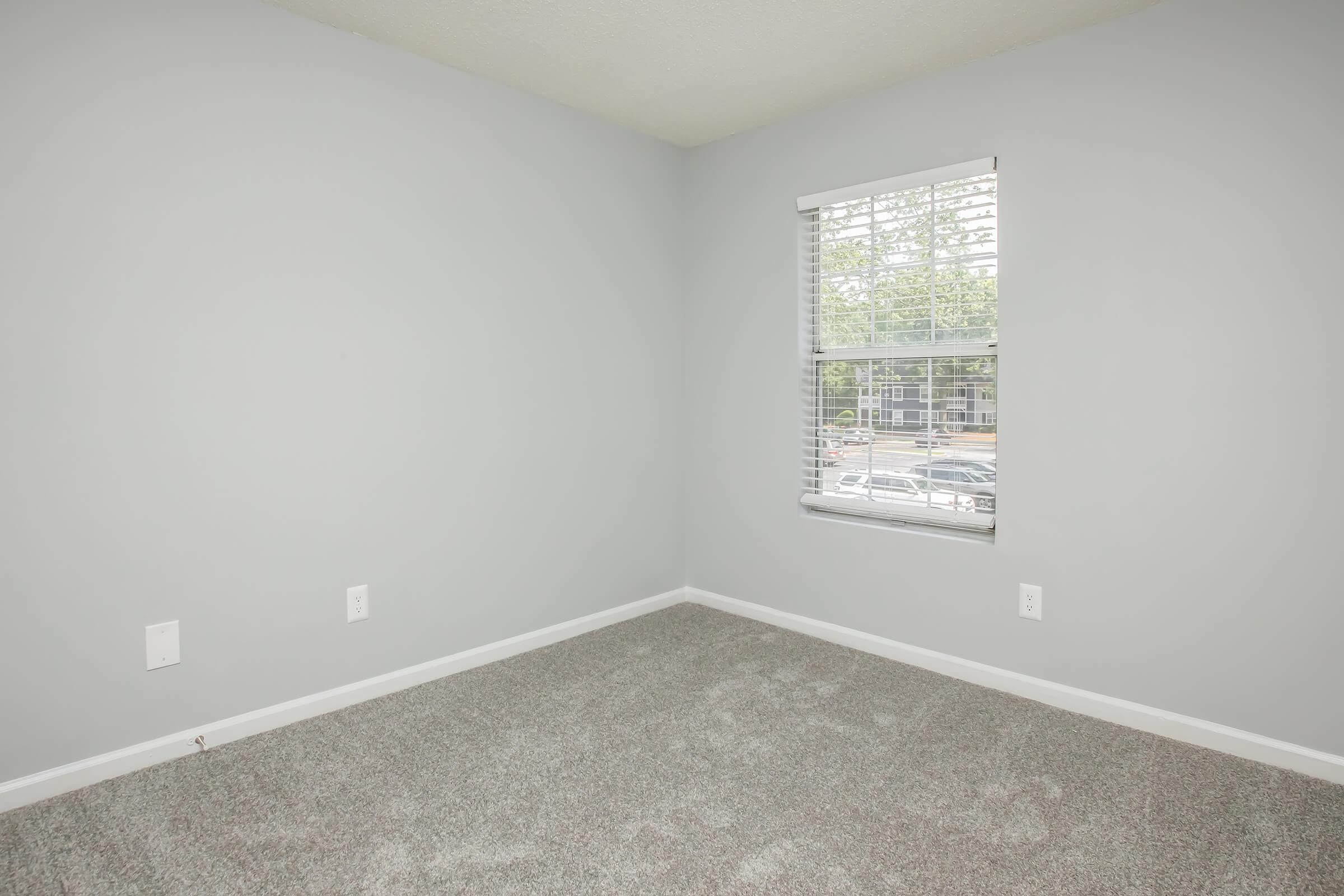 Empty room with light gray walls and carpeted flooring. A window with white blinds lets in natural light, revealing a view of a parking area outside. The corner of the room is slightly visible, with electrical outlets on the wall.