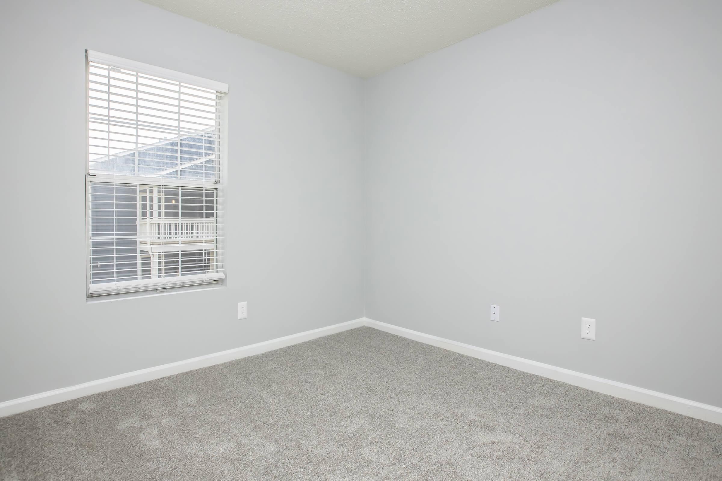 An empty room with light gray walls and carpeted floor. A window with white blinds lets in natural light, revealing a balcony outside. The room has no furniture, creating a spacious and minimalist atmosphere. There are electrical outlets on the walls.