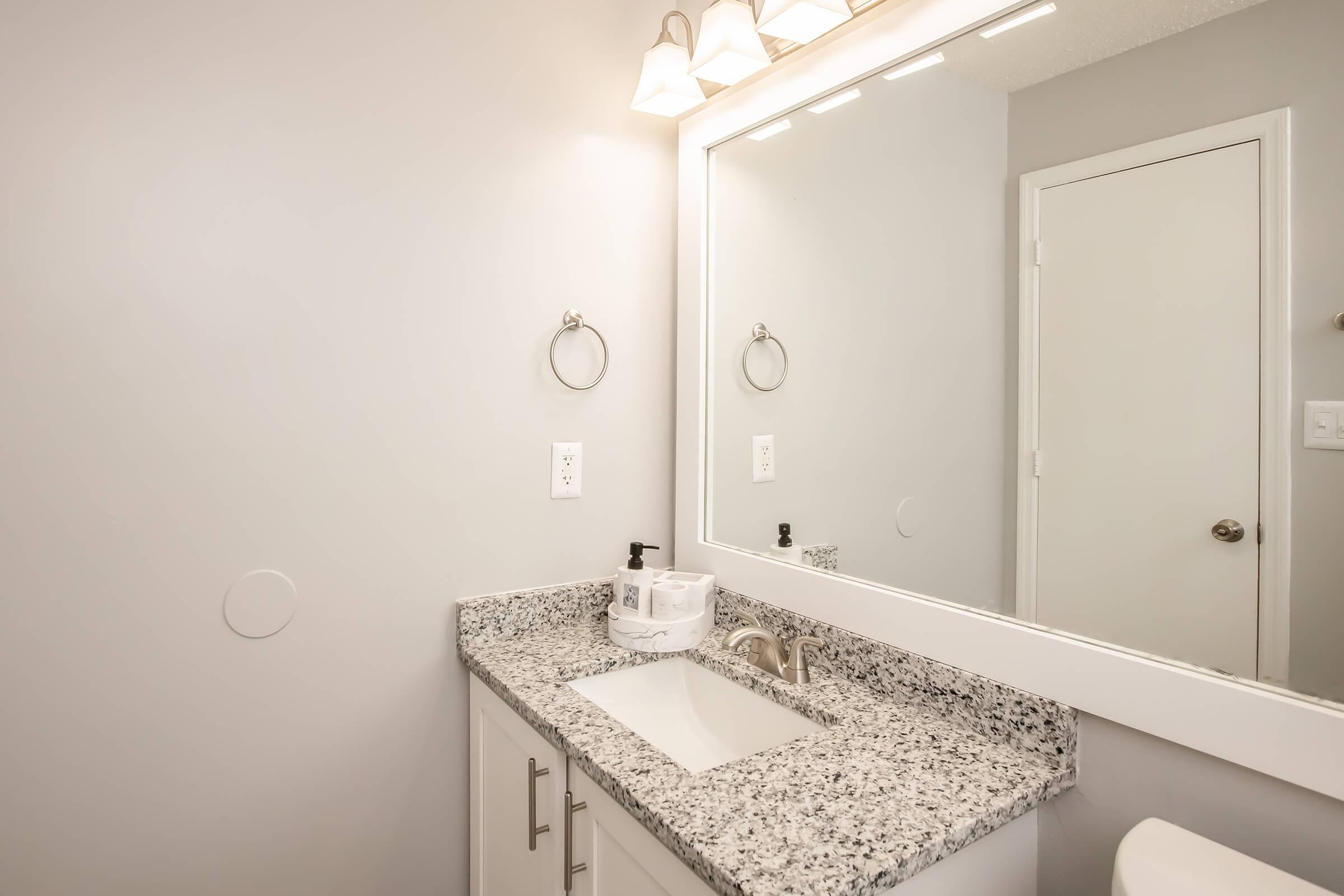 Modern bathroom interior featuring a granite countertop with a sink, a large wall mirror, and light fixtures above. The walls are painted in a light color, and a white door is visible in the background. A towel ring is mounted on the wall beside the sink.