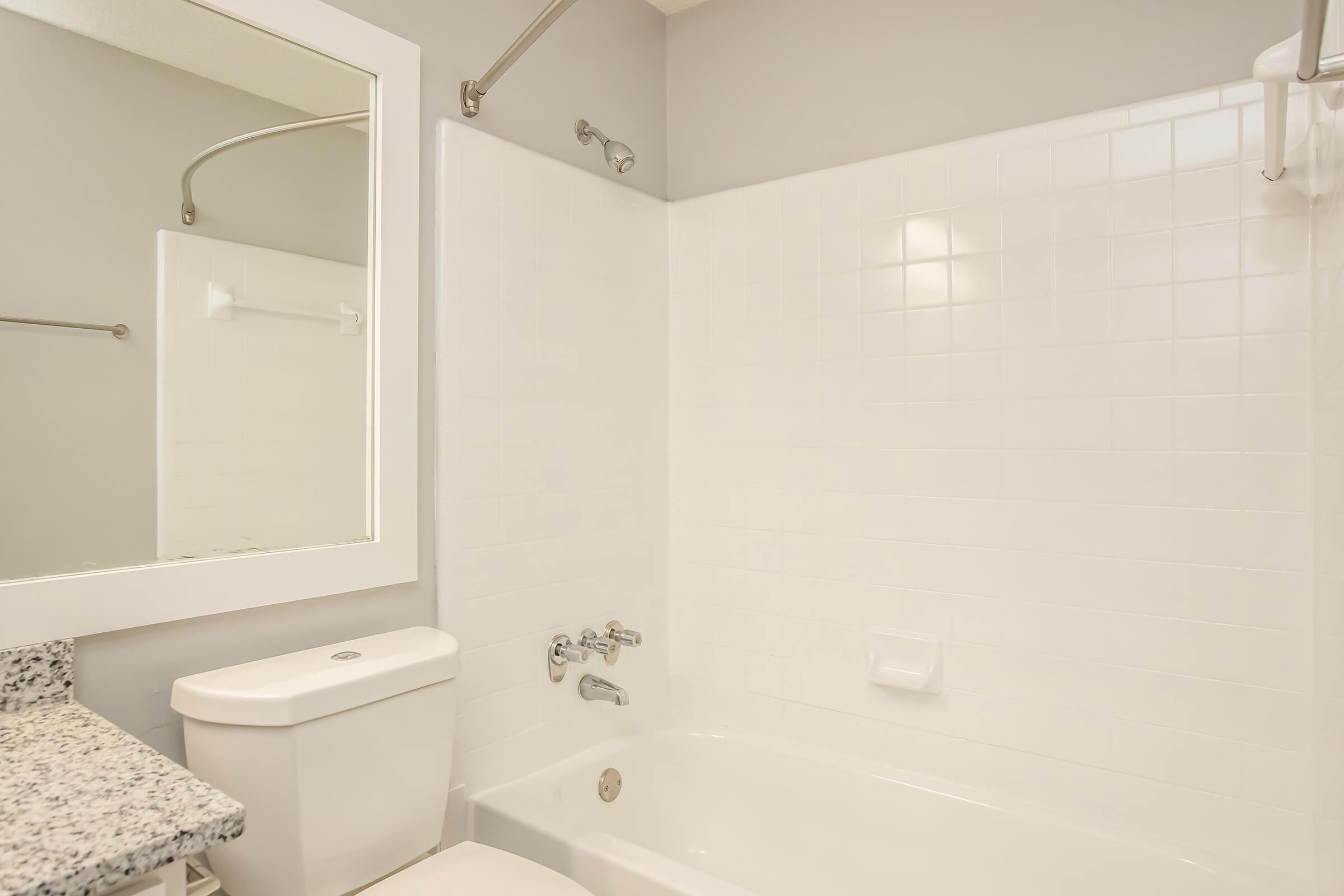 A clean bathroom featuring a white tiled shower and bathtub, a granite countertop with a sink, and a large mirror above the sink. The walls are painted light grey, and there are towel bars and a shower curtain rod installed. The overall appearance is modern and tidy.