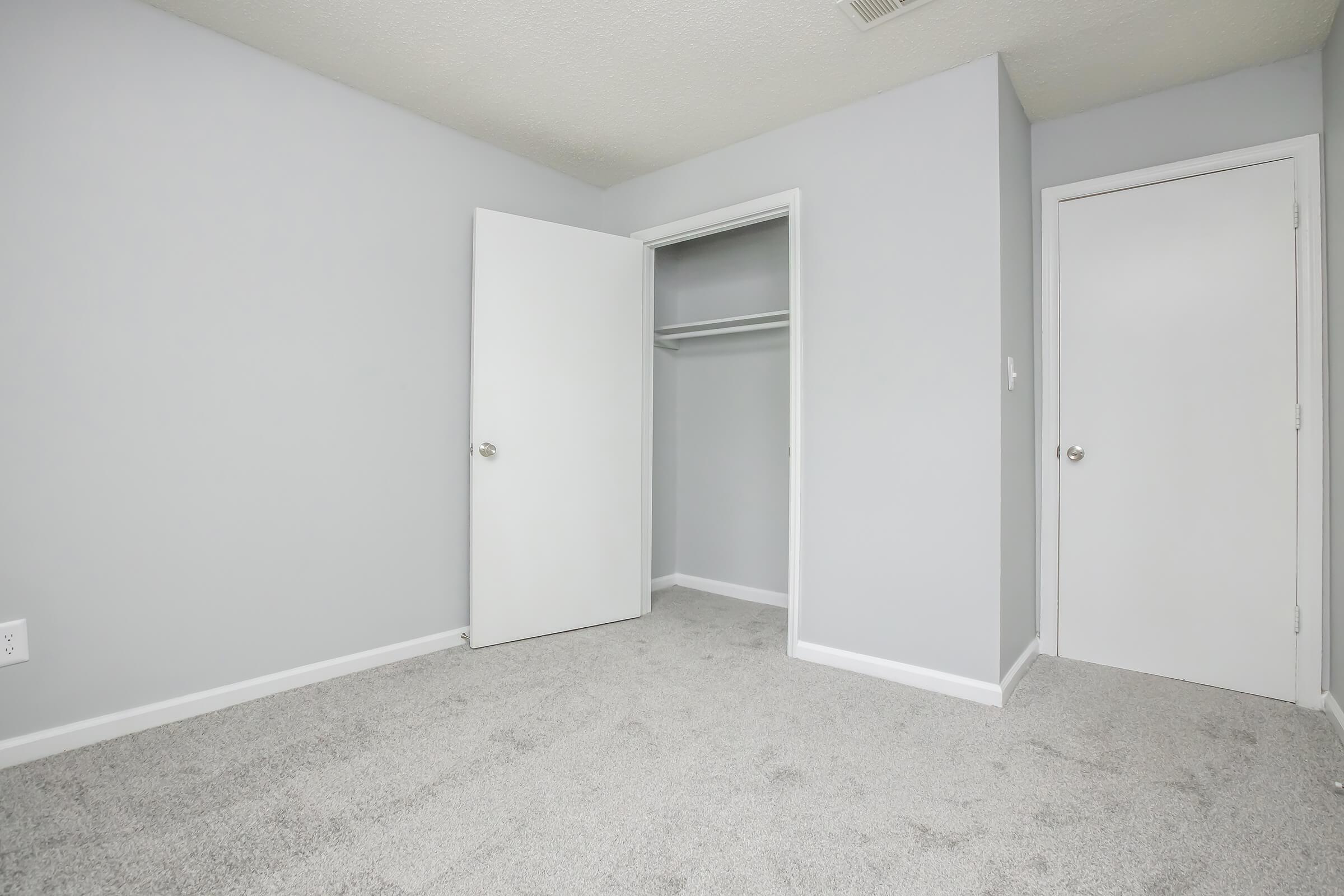 A simple, empty bedroom featuring light gray walls and carpet. There are two white doors: one leading to a closet and another to an adjacent room. The space appears well-lit and ready for furniture.