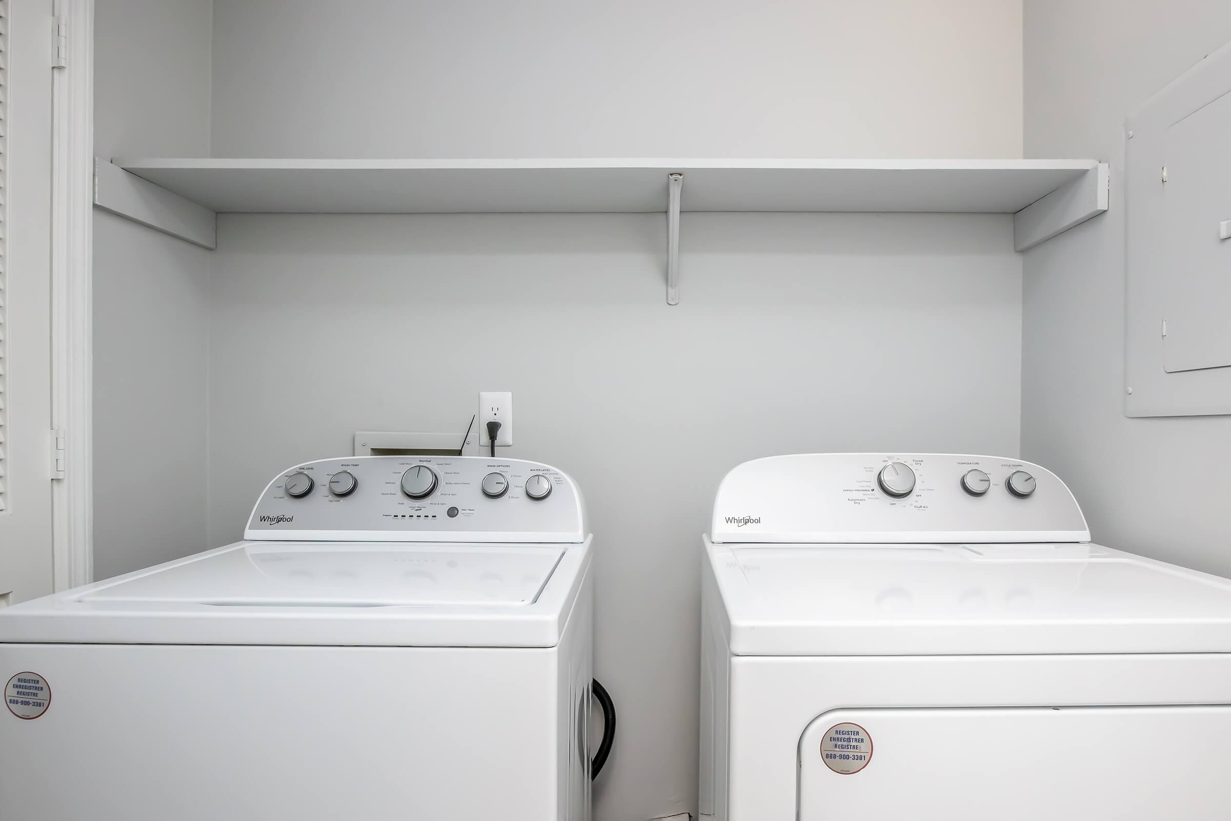 Two white washing machines side by side in a laundry room. A simple shelf is mounted above them against a light gray wall. The machines have control knobs and settings visible on their fronts, with a power outlet nearby.