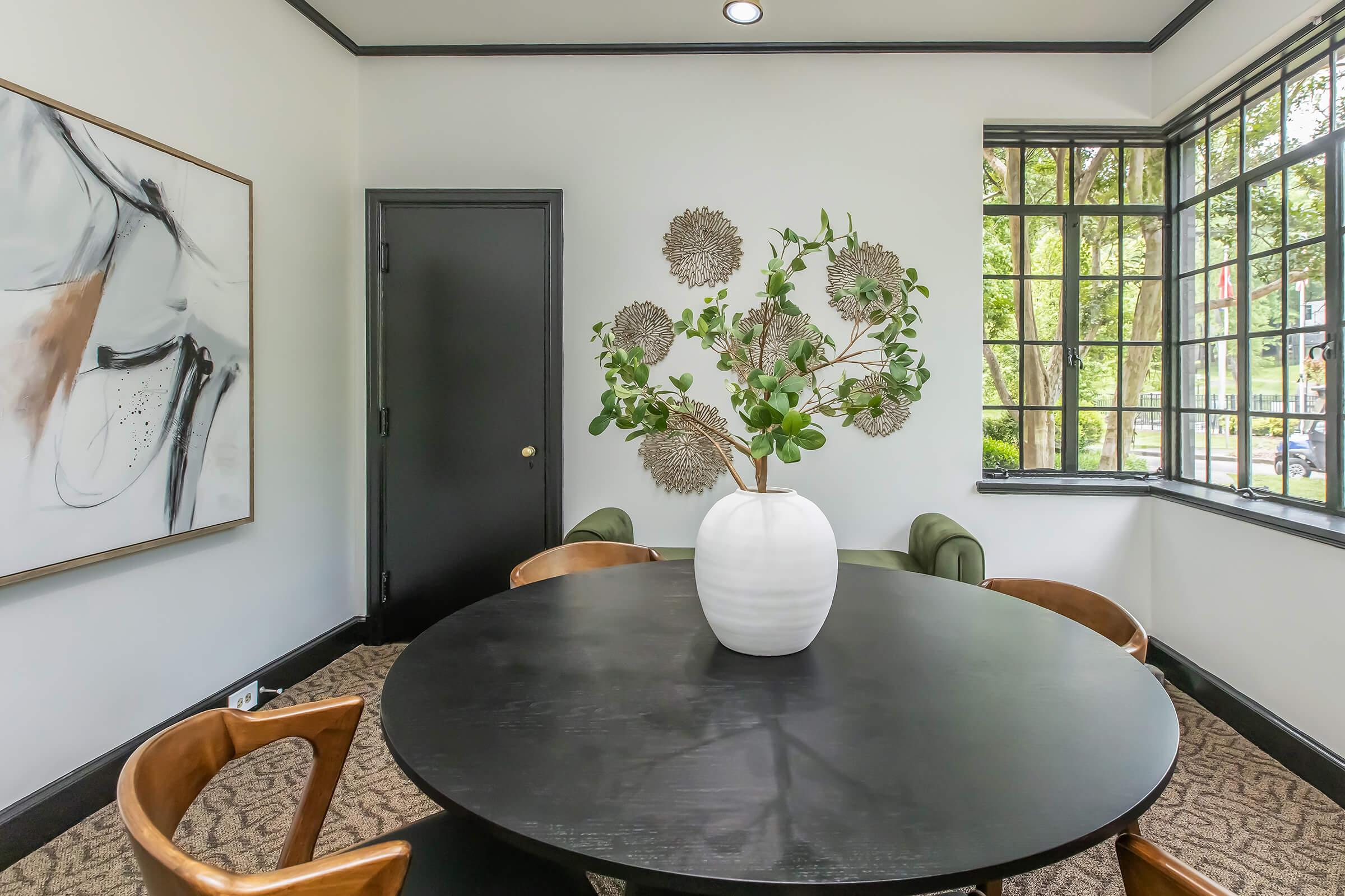A modern dining area featuring a round black table surrounded by wooden chairs. In the center, a large white vase holds a leafy plant. The walls are adorned with a contemporary artwork, and large windows provide natural light, highlighting the minimalist decor.