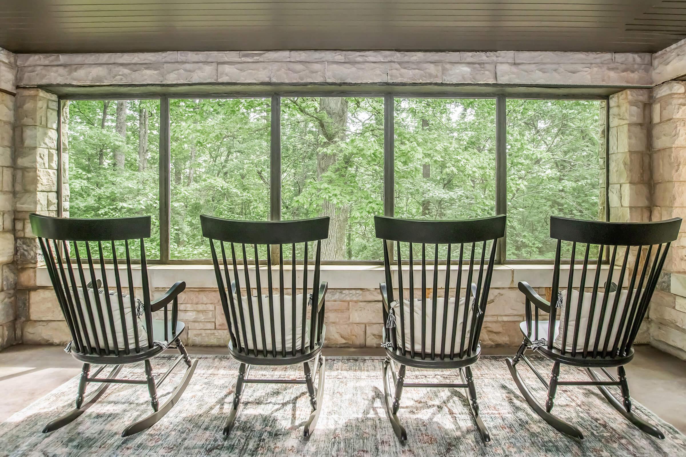 Four black rocking chairs arranged in a row facing large windows that overlook a lush green forest. The room features stone walls and a cozy ambiance, creating a peaceful atmosphere ideal for relaxation.