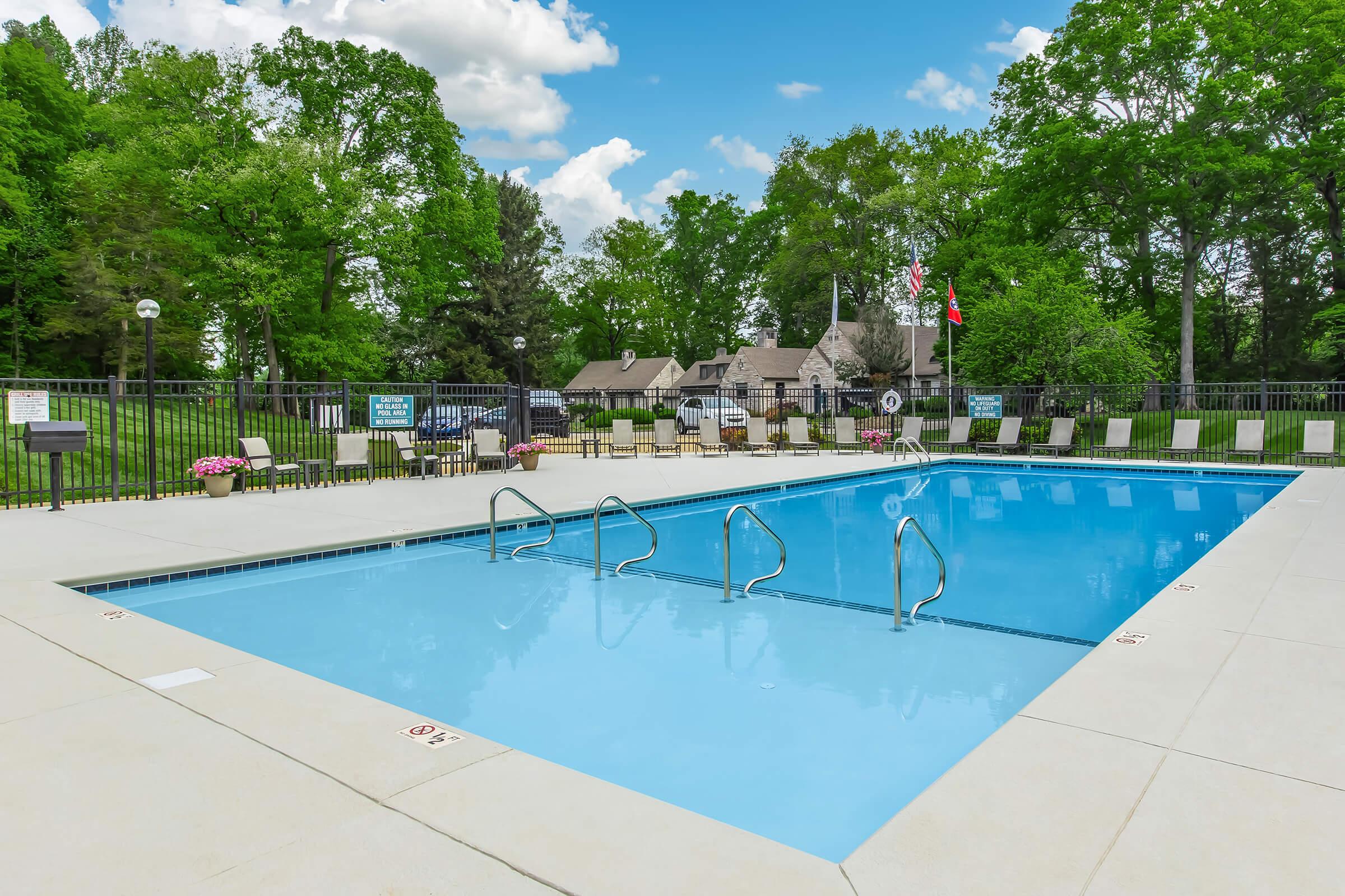 A clean outdoor swimming pool with step rails, surrounded by a concrete deck. Lounge chairs line one side, and lush green trees and grass provide a natural backdrop. A clubhouse with a traditional design is visible in the background, along with decorative flowers and safety signage. The sky is bright with fluffy clouds.