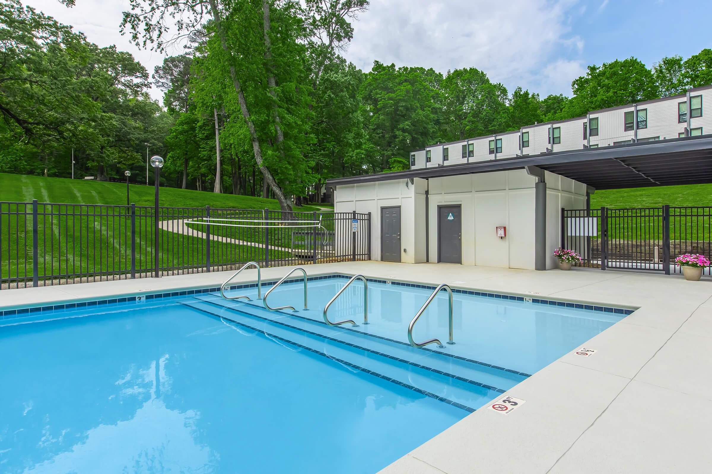 A clear swimming pool with steps leading in, surrounded by a tiled deck. Lush green trees and grass are in the background, along with a grassy area and a building partially visible to the right. The pool area is well-maintained, featuring fencing and outdoor lighting for ambiance.