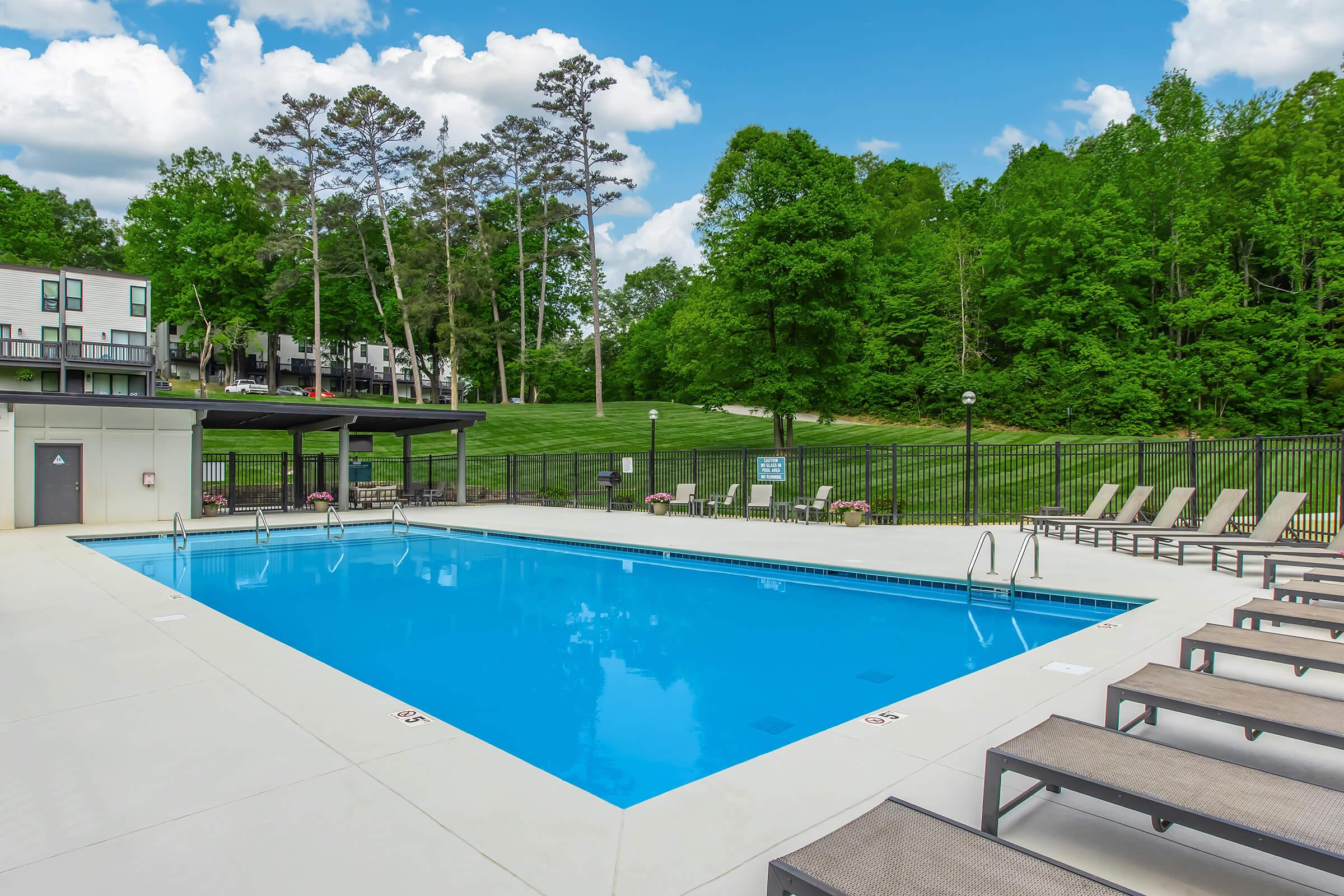 A clear blue swimming pool surrounded by lounge chairs, set against a backdrop of lush green trees and a bright sky with white clouds. The pool area is neatly maintained, with a fence in the background and a grassy hill visible beyond the trees.