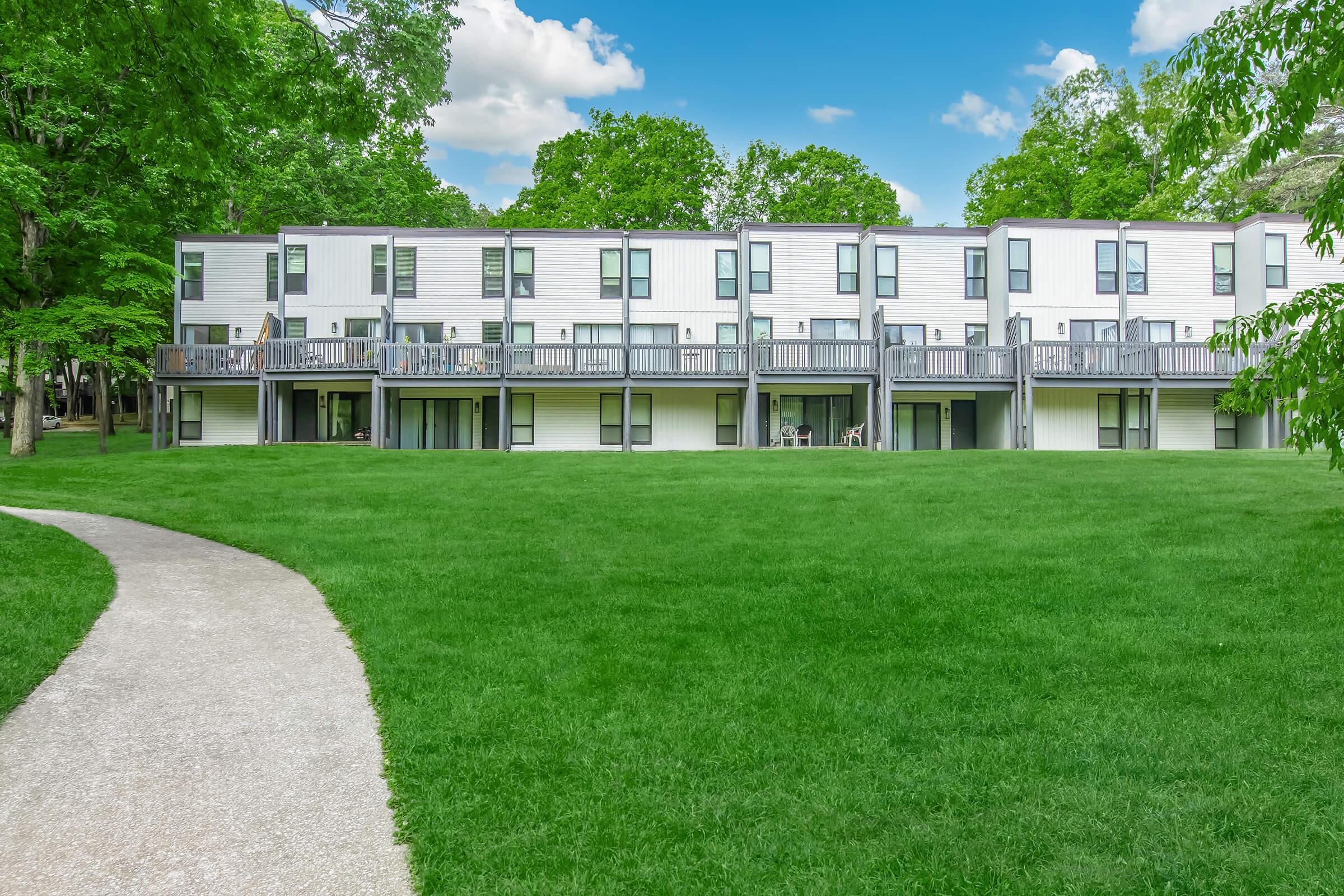 A modern multi-unit residential building with white siding and balconies, surrounded by lush green grass and trees. A winding pathway leads up to the building, set against a clear blue sky with a few fluffy clouds.
