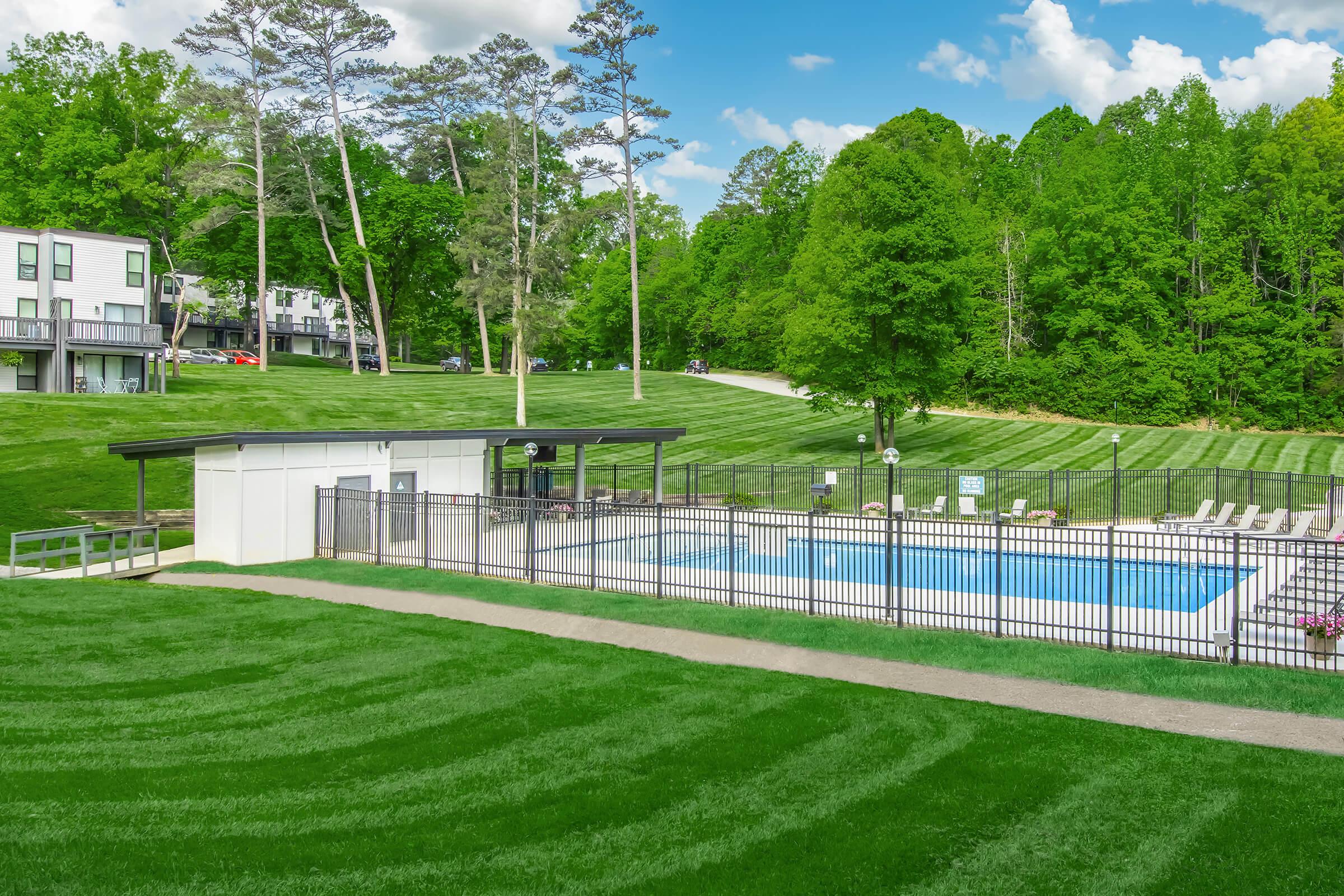 A well-maintained outdoor swimming pool surrounded by a manicured lawn, with green trees in the background. The pool area features a fenced perimeter, lounge chairs, and a pool house. Clear blue skies and fluffy clouds add to the inviting atmosphere of the space.