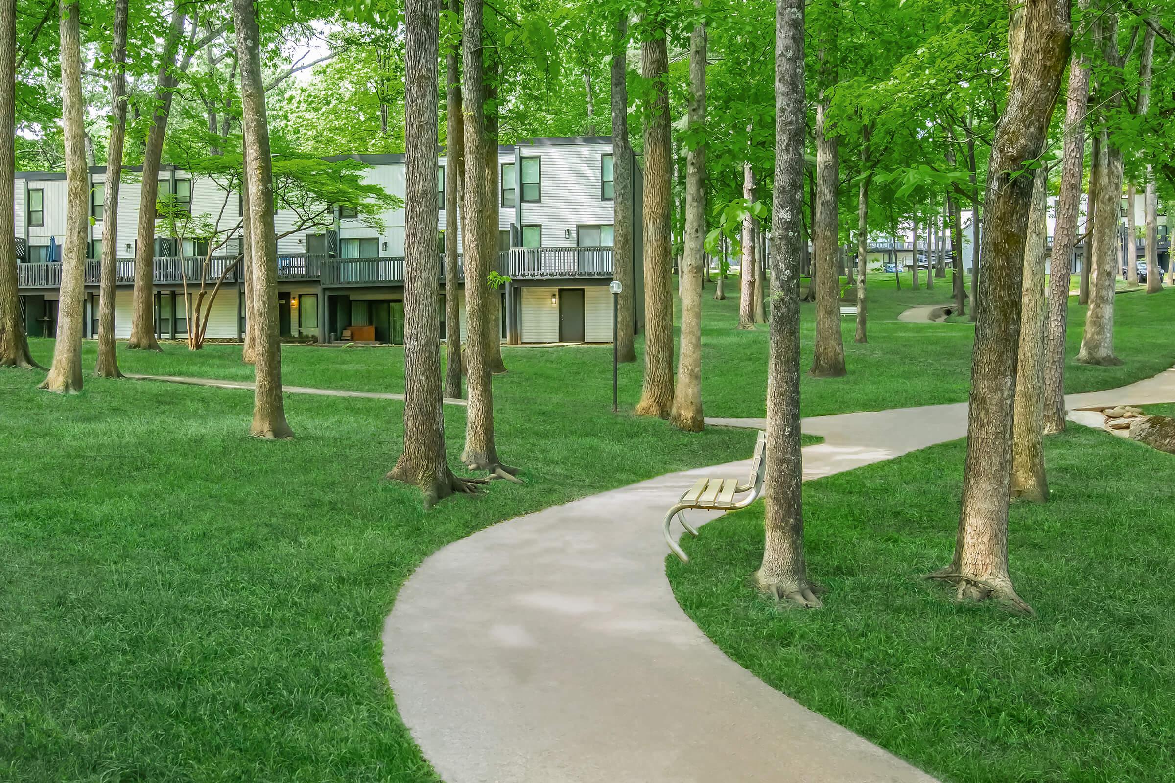 A serene outdoor scene featuring a winding path through a lush green park. Tall trees frame the path, leading to a cluster of modern, multi-story buildings with balconies. The grass is well-maintained, creating a peaceful atmosphere ideal for walking or relaxing in nature.