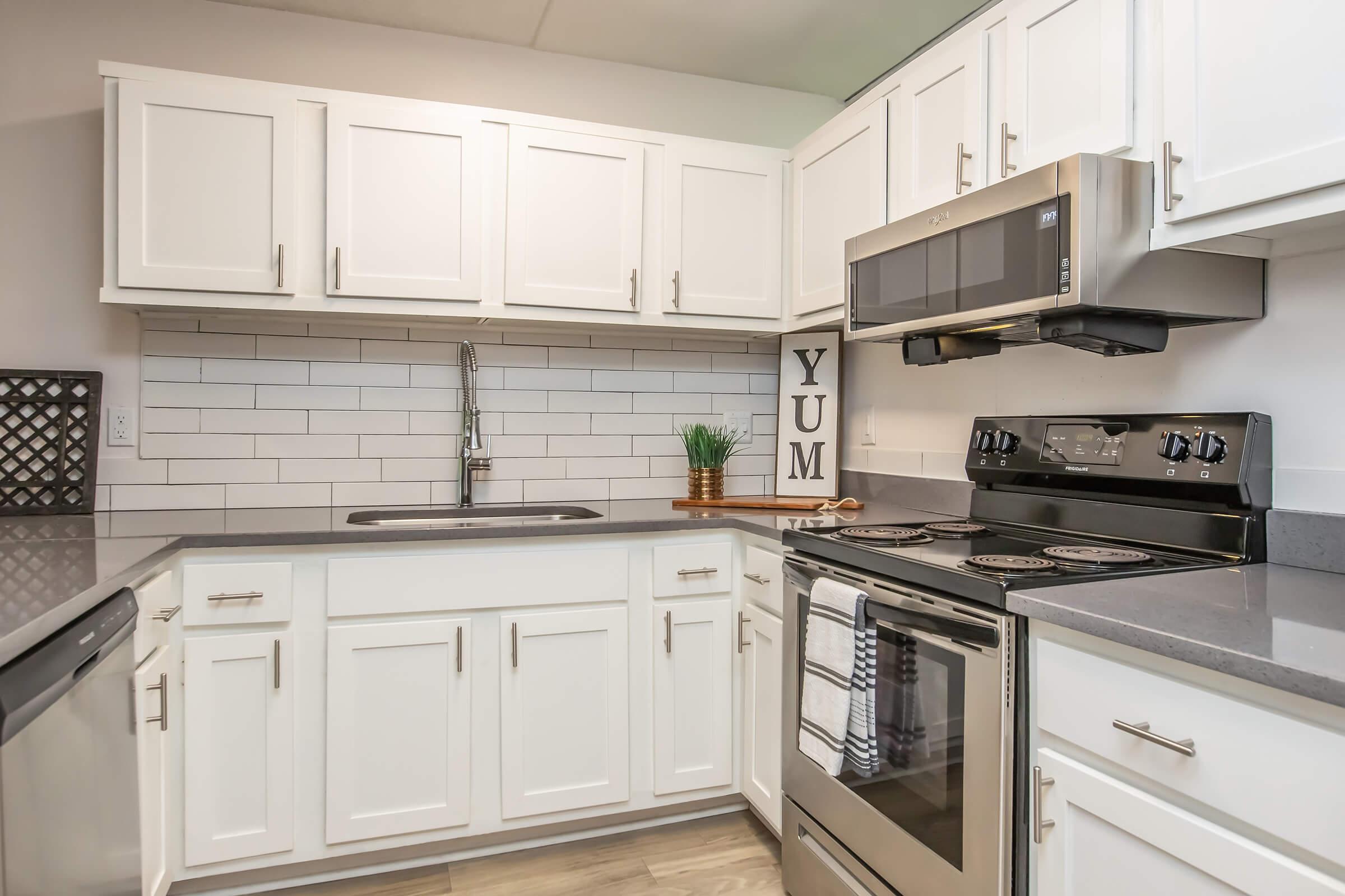Modern kitchen with white cabinets and a sleek countertop. Features stainless steel appliances, including a microwave and oven. The backsplash consists of white tiles, and there’s a small potted plant. A decorative sign with the word "YUM" is visible on the countertop, adding a touch of warmth to the space.