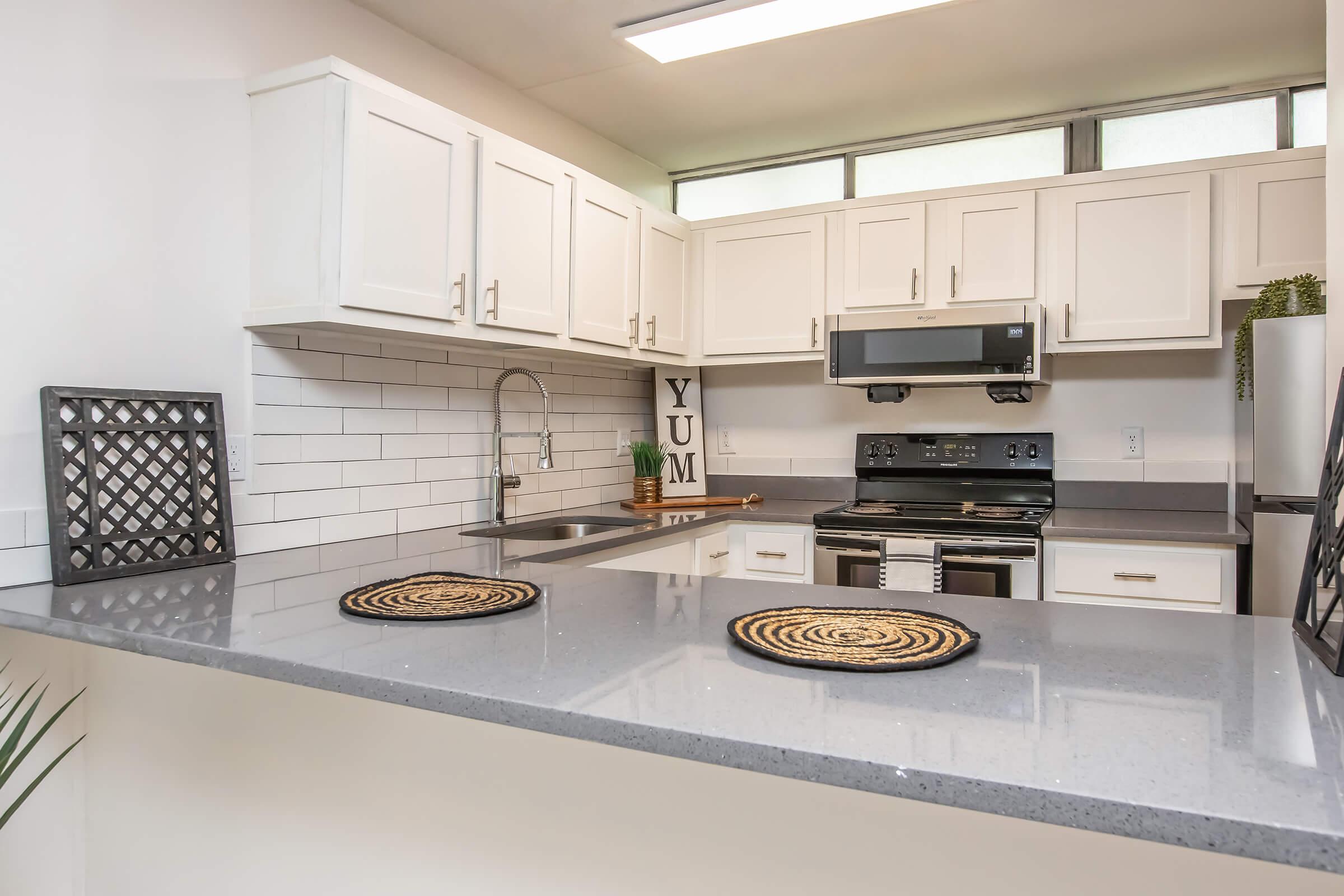A modern kitchen featuring white cabinets, a gray countertop, and stainless steel appliances. The sink is positioned under a window, and decorative items like woven placemats and a sign that reads "YUM" add a personal touch. The space is well-lit and organized, conveying a clean and inviting atmosphere.