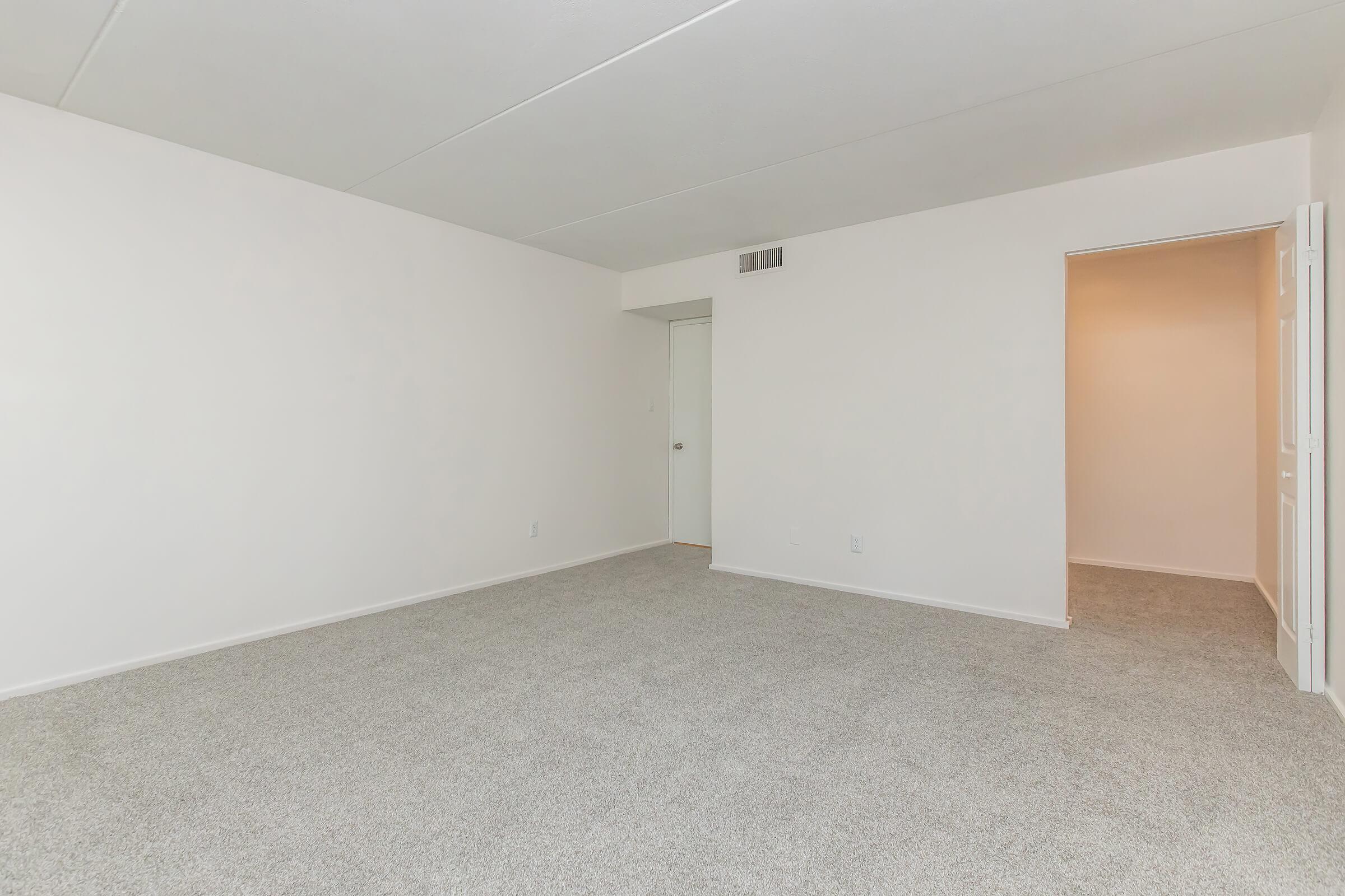 Empty room with light beige carpet and plain white walls. A single door leads to another room, and there are no furnishings or decor, creating a spacious and minimalistic atmosphere. Natural light may illuminate the space, highlighting the clean lines and simplicity of the design.