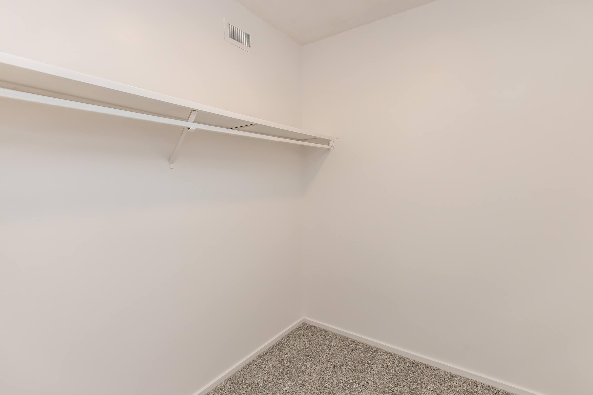 Empty closet interior with a white wall and a single shelf mounted on one side. The floor is covered with light-colored carpeting. There are no items or furniture present, giving the space a clean and minimalistic appearance.