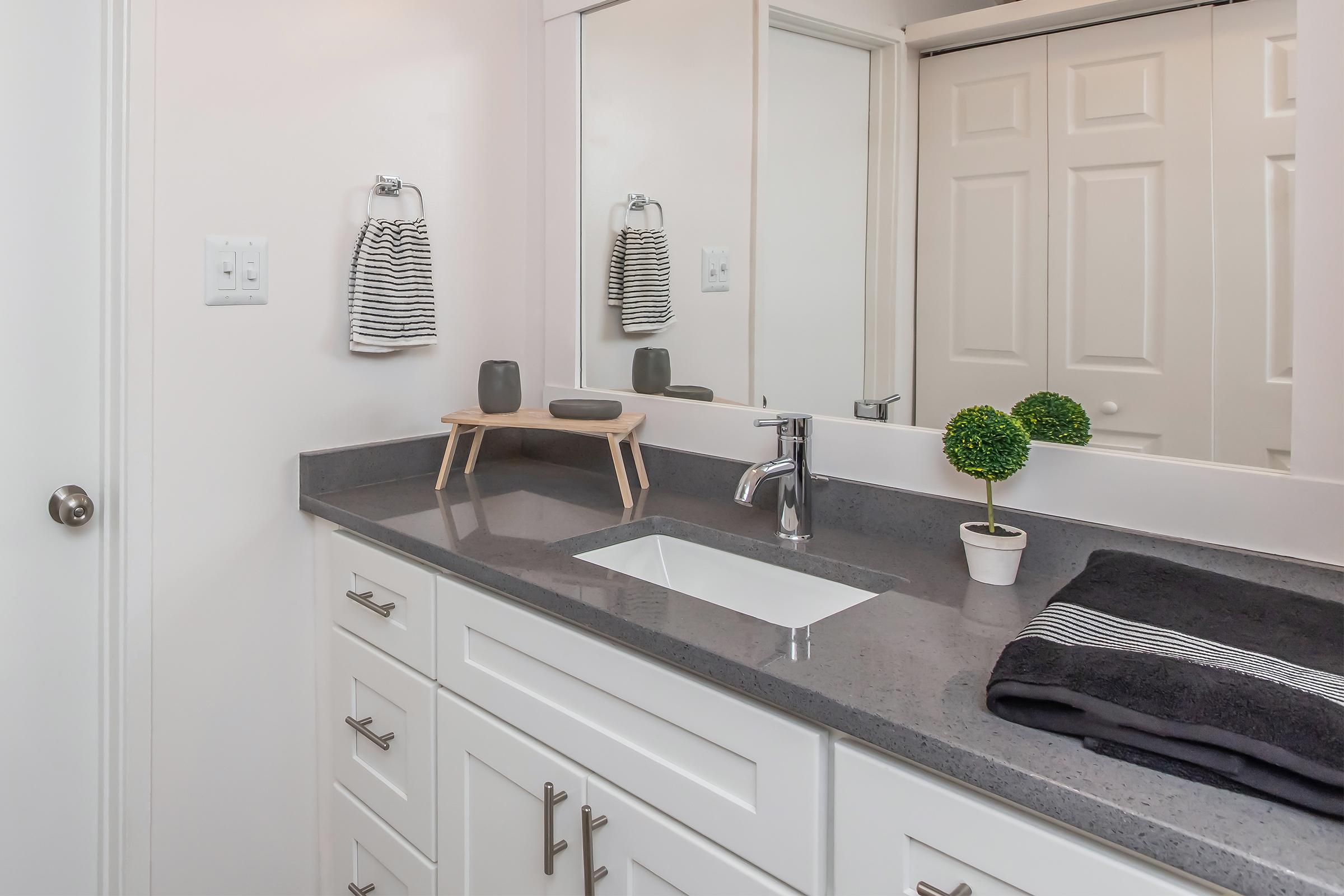 A modern bathroom countertop featuring a sleek gray surface, a rectangular sink, and minimalist decor. There's a wooden stool holding a small decorative item, and two potted plants. White cabinetry with stylish handles complements the light-colored walls, while bathroom accessories include striped towels and matching hand towels.