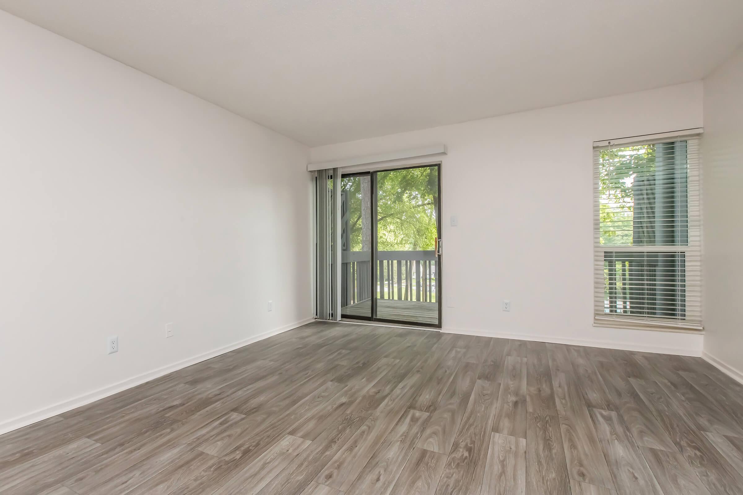 A brightly lit empty living room with light-colored walls and wood-like flooring. There is a sliding glass door leading to a small balcony, flanked by a window with blinds. The space is clean and uncluttered, suitable for decorating.