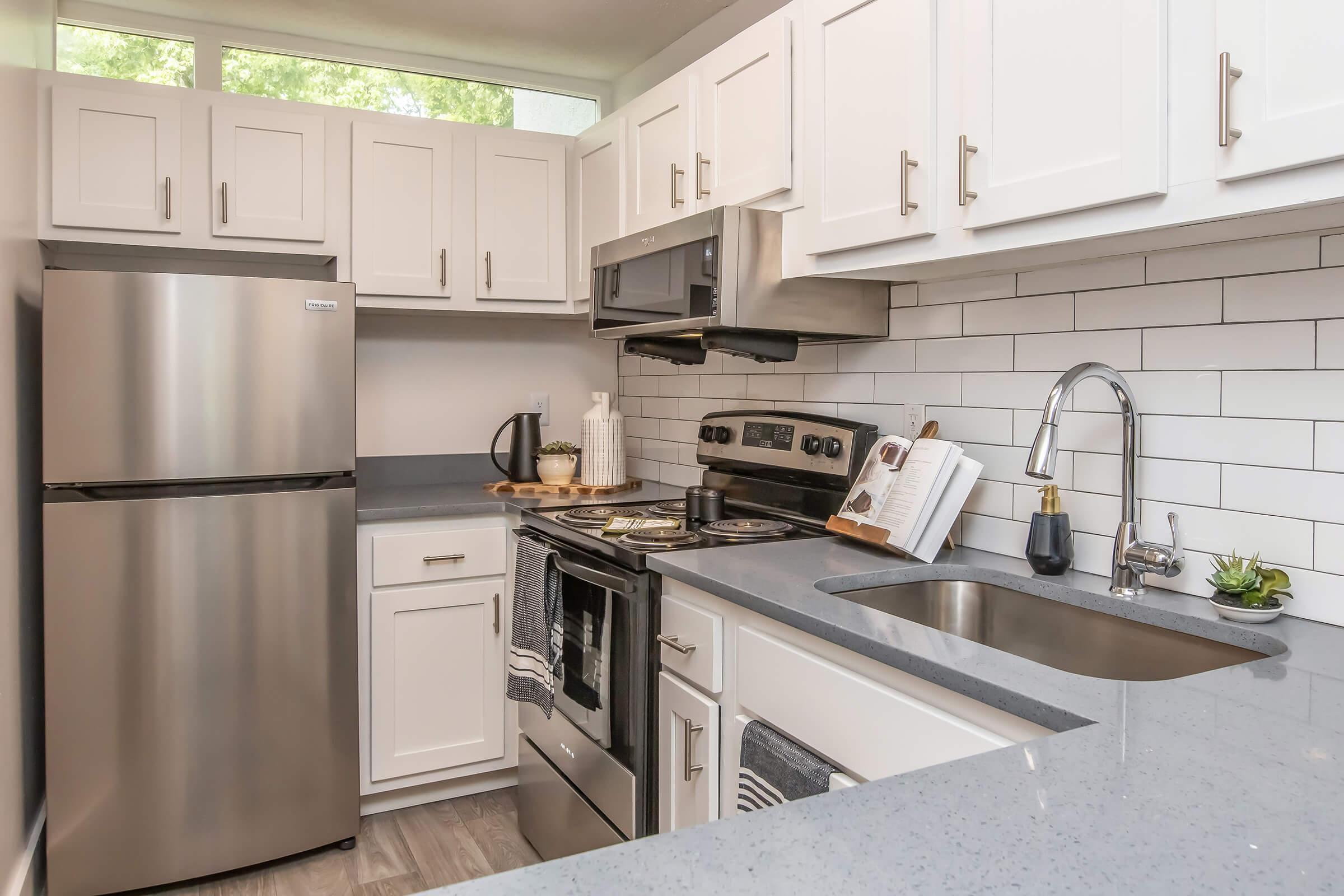 Modern kitchen featuring white cabinetry, stainless steel appliances including a refrigerator, stove, and microwave. Countertops are gray with a sink, and there is a cookbook and decorative items on the counter. The backsplash consists of white tiles, and the area is well-lit with natural light.