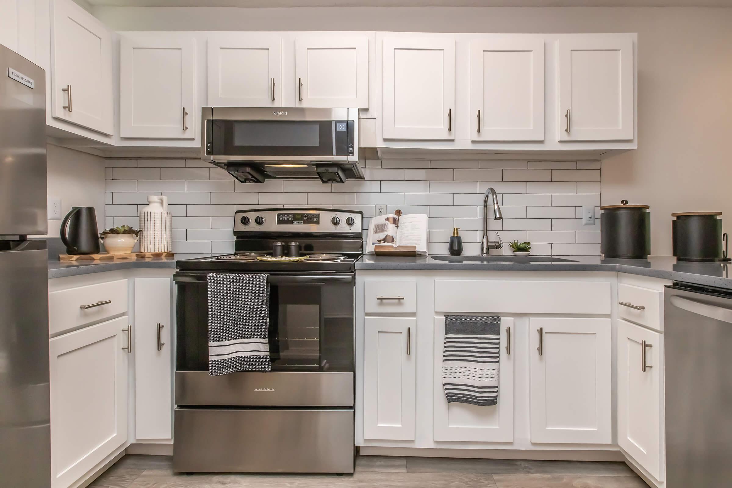 A modern kitchen with white cabinetry, stainless steel appliances, and a gray countertop. An oven is centered with a stovetop, flanked by a microwave above. Decorative items like a plant, dish towels, and utensils are arranged neatly, and the backsplash features white subway tiles.