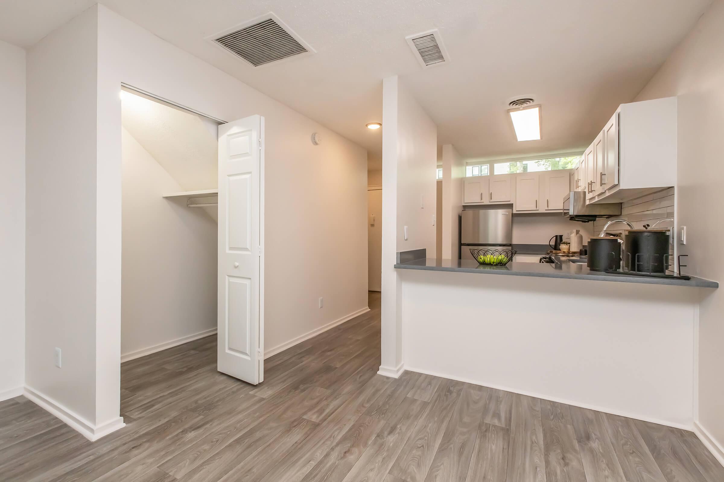 A modern kitchen and living area featuring light-colored walls, gray wood-like flooring, and a breakfast bar. The kitchen includes white cabinets, stainless steel appliances, and a glimpse of a closet space through an open door. Natural light fills the room from a nearby window.